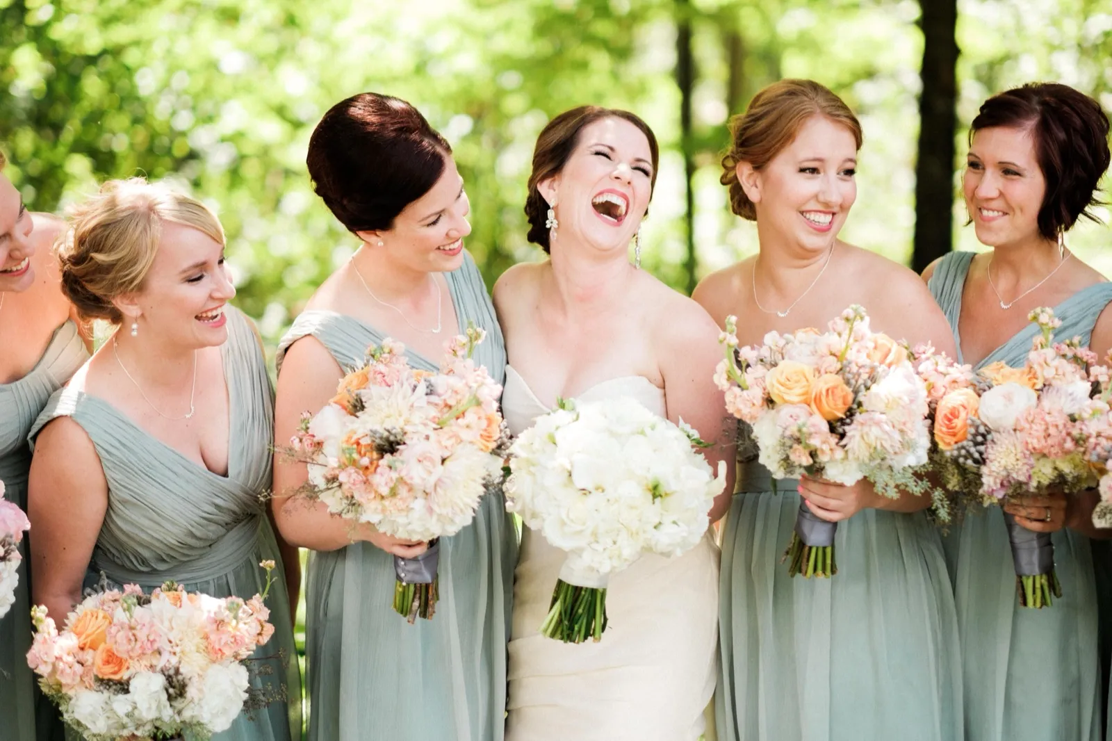 The bride laughs open-mouthed while her bridesmaids in sage dresses smile around her, all holding peach and ivory bouquets in a sunlit woods. — Tim Larsen Photography, Brainerd Lakes MN