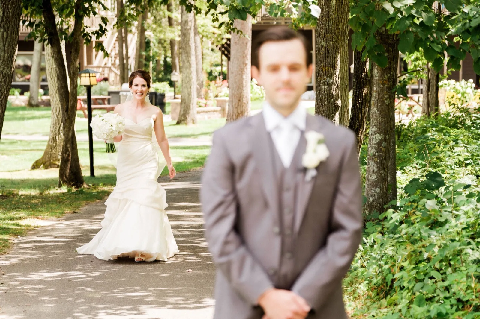 The bride walks toward her groom, who waits with his back turned in the foreground, on a tree-lined path at Grand View Lodge. — Tim Larsen Photography, Brainerd Lakes MN