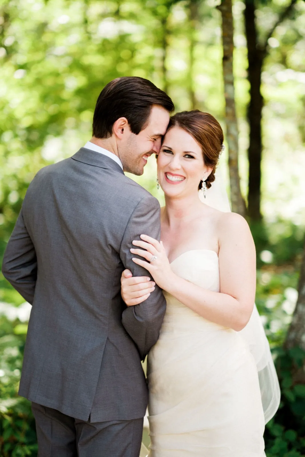 The bride smiles toward the camera as the groom leans in to nuzzle her cheek, surrounded by soft green forest at Grand View Lodge. — Tim Larsen Photography, Brainerd Lakes MN