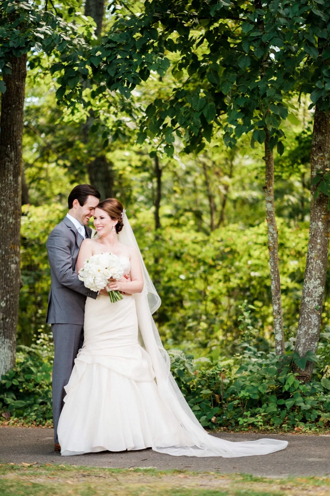 The groom rests his forehead against the bride's temple as she smiles and holds a white bouquet, the two standing together beneath a wide canopy of summer trees with her veil and gown trailing across the path. — Tim Larsen Photography, Brainerd Lakes MN