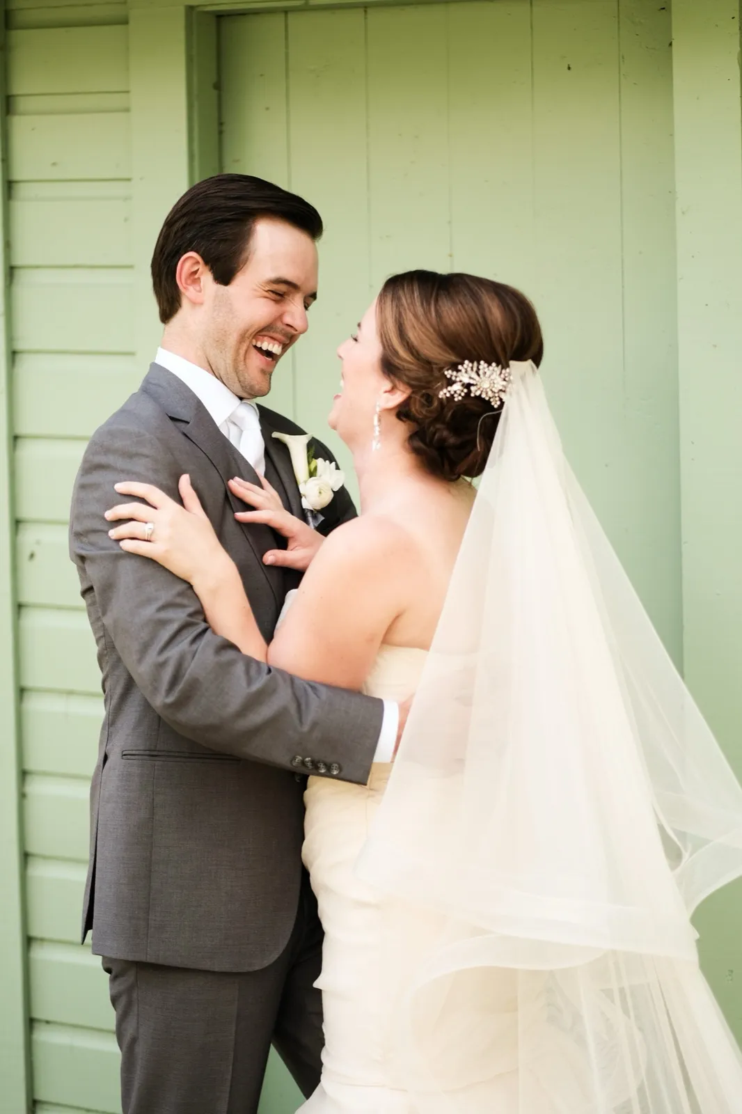 The bride and groom laugh together face-to-face against a mint-green clapboard wall, the bride's long veil draping to one side. — Tim Larsen Photography, Brainerd Lakes MN