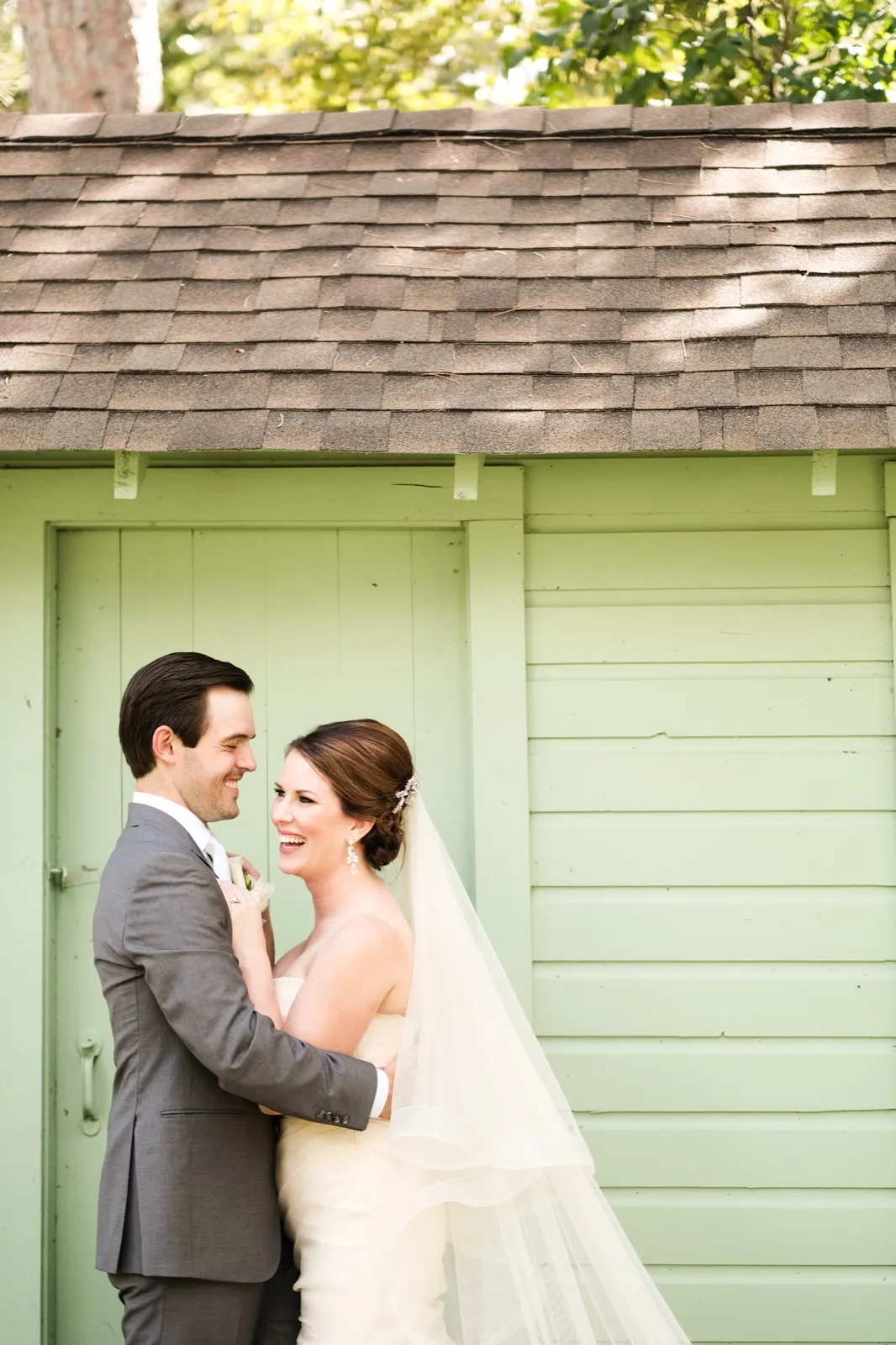 The bride laughs while facing her groom outside a mint-green cottage with a shingled roof, her veil trailing across the frame. — Tim Larsen Photography, Brainerd Lakes MN