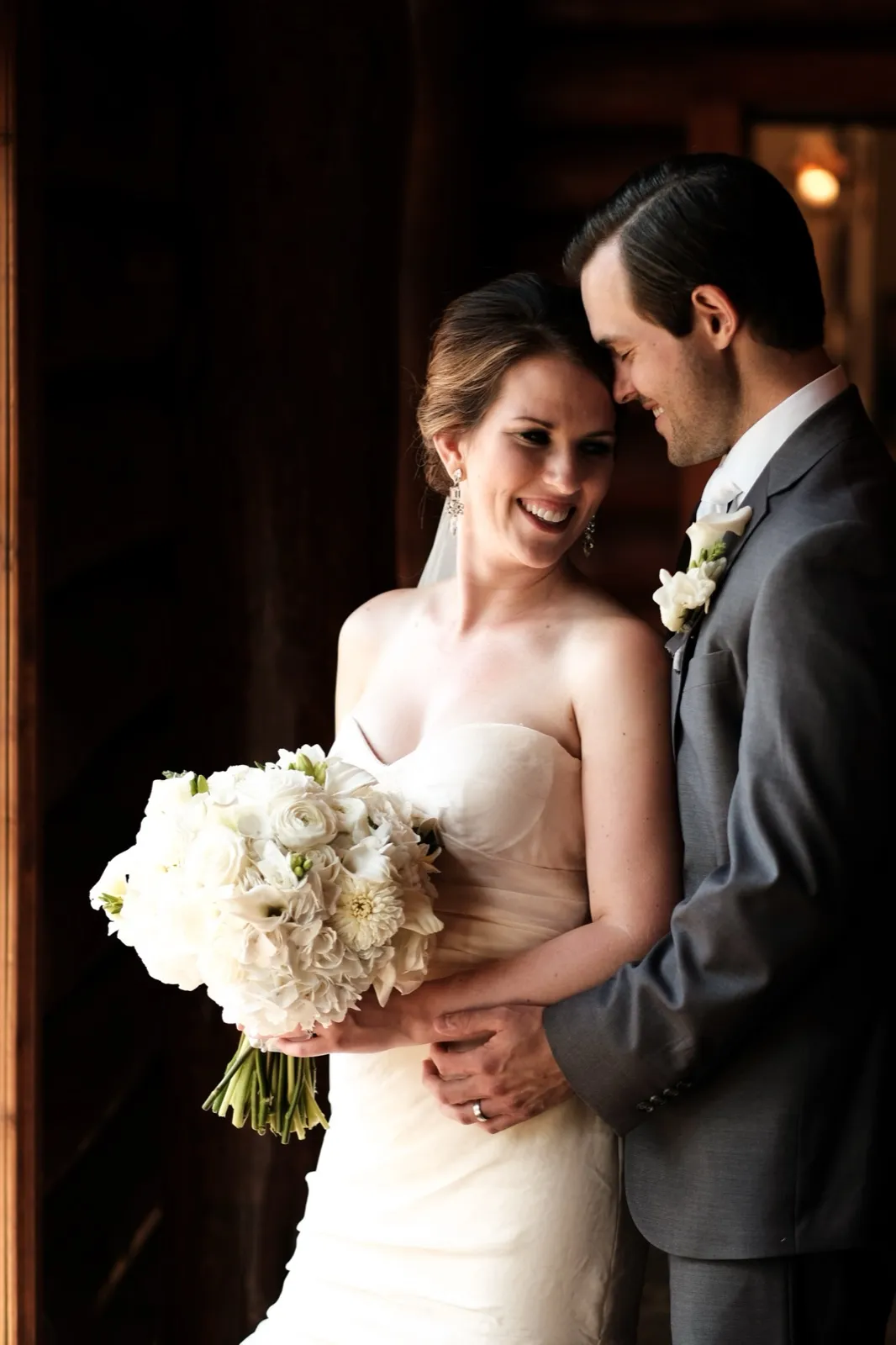 The bride smiles softly while holding a white bouquet as the groom leans his forehead against hers in a dim wood-paneled doorway lit by window light. — Tim Larsen Photography, Brainerd Lakes MN
