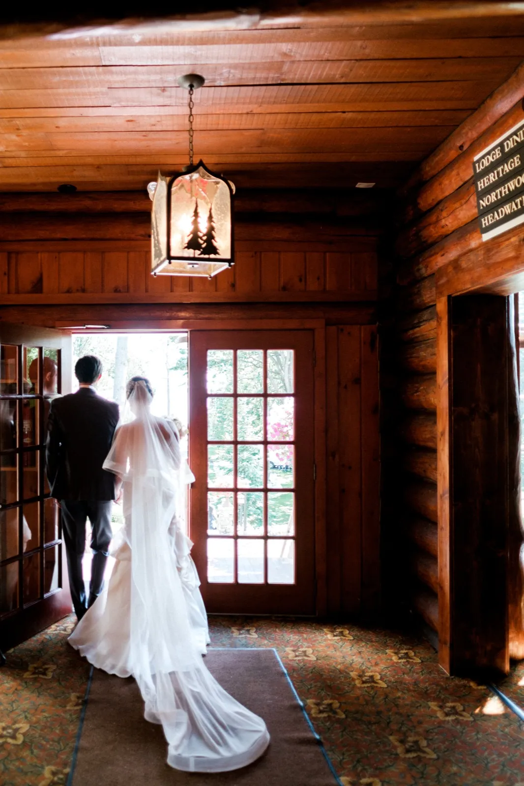 The bride's veil and train trail behind her as she walks with the groom toward a sunlit French door beneath a pine-tree lantern in the wood-paneled lodge entryway. — Tim Larsen Photography, Brainerd Lakes MN