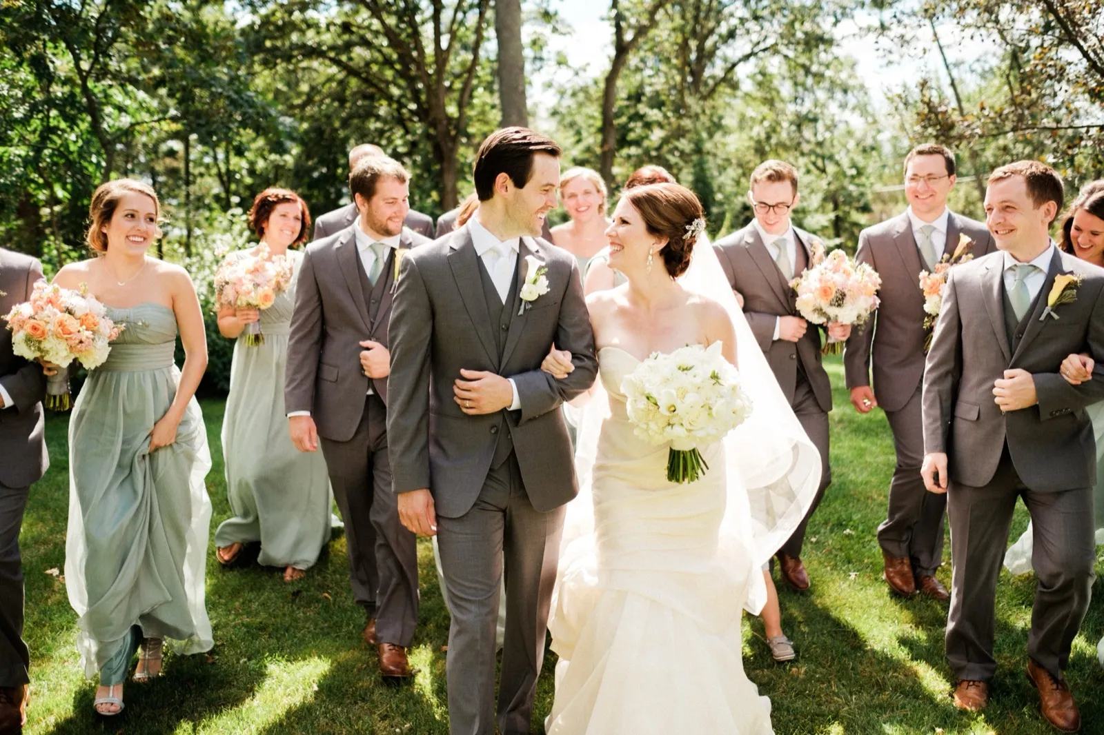 The bride and groom walk arm-in-arm across a sunlit lawn with their bridal party flanking them on both sides, all laughing together. — Tim Larsen Photography, Brainerd Lakes MN