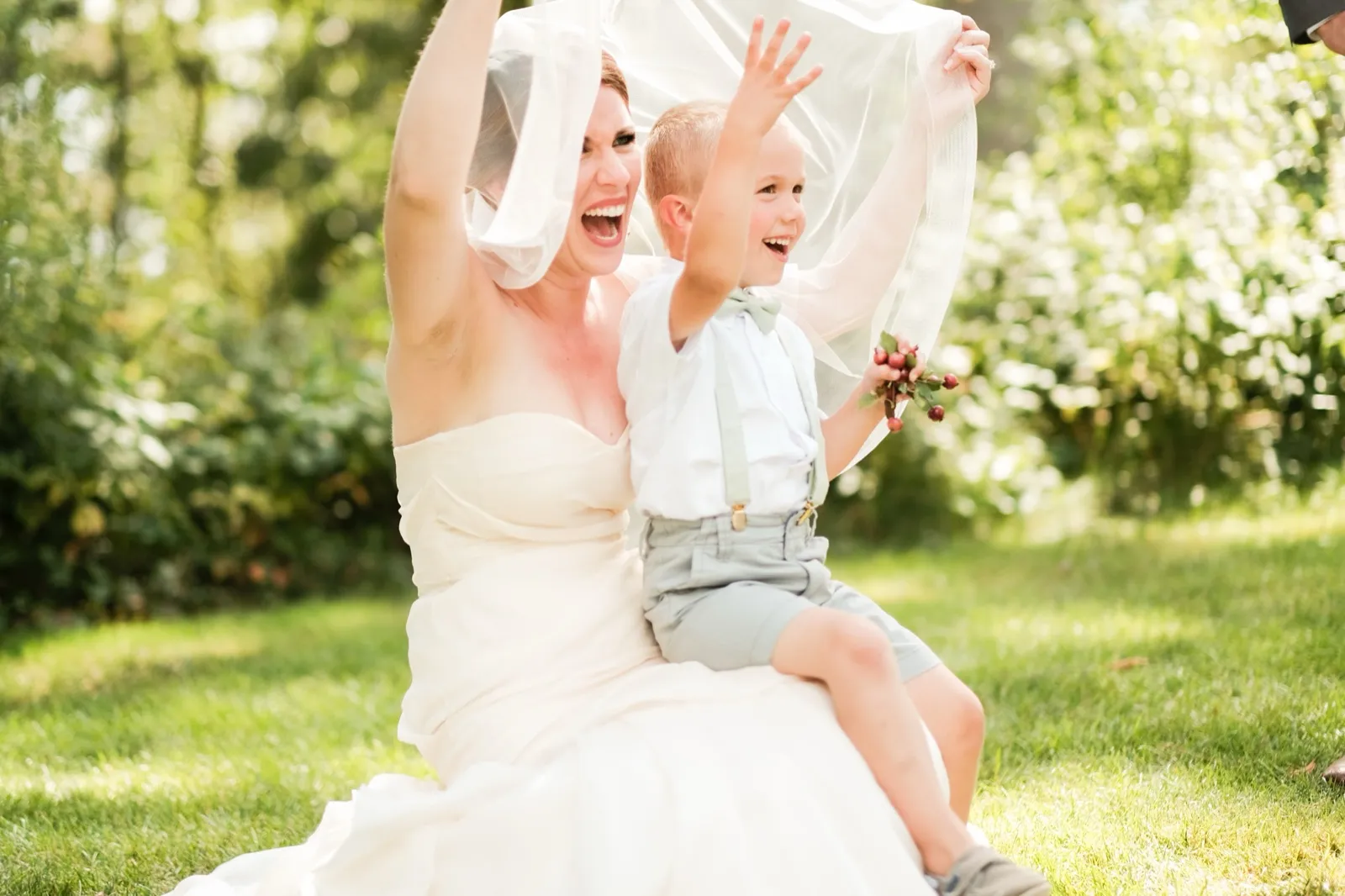The bride and a young ring bearer throw their arms up and laugh together under a sunlit veil held overhead on a garden lawn. — Tim Larsen Photography, Brainerd Lakes MN