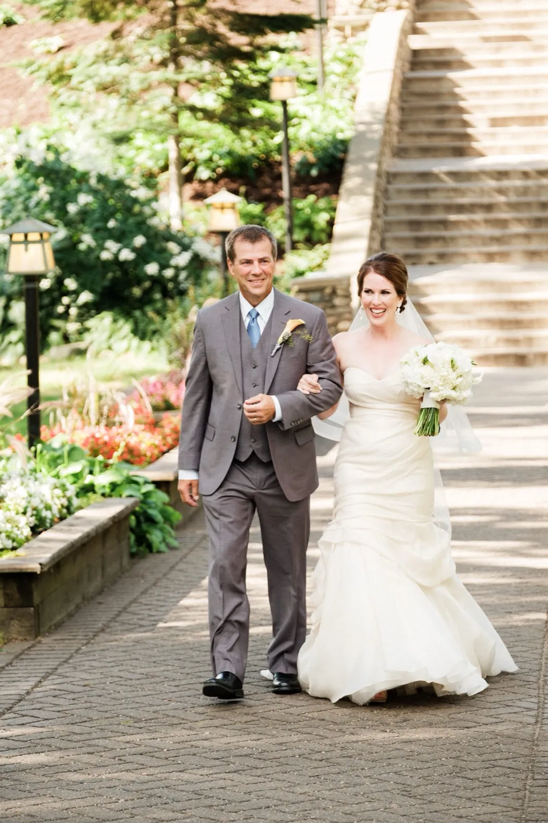 The bride walks arm-in-arm with her father down a stone path past flower beds and lamp posts, with Grand View Lodge's stairway rising behind them. — Tim Larsen Photography, Brainerd Lakes MN