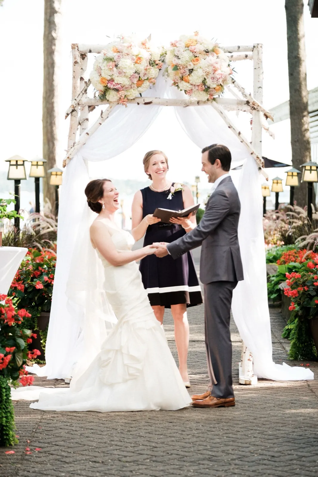 The bride laughs while holding her groom's hands beneath a birch and floral ceremony arch as the officiant smiles between them at a lakeside Grand View Lodge ceremony. — Tim Larsen Photography, Brainerd Lakes MN