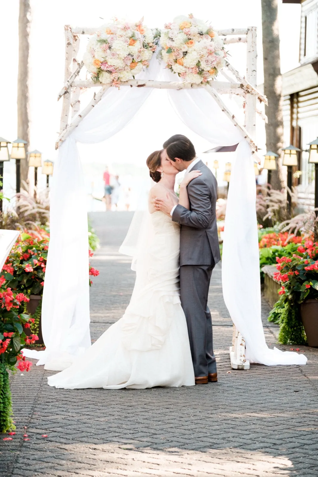 The bride and groom share a kiss beneath a birch ceremony arch draped with white fabric and topped with blush and ivory florals, flanked by red flower beds and lantern posts on a brick walkway. — Tim Larsen Photography, Brainerd Lakes MN