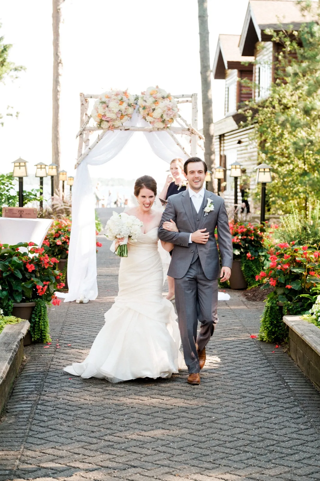 A bride and groom walk arm-in-arm down a brick aisle after their ceremony, beaming, with a birch-and-floral arch and the Grand View Lodge buildings behind them. — Tim Larsen Photography, Brainerd Lakes MN