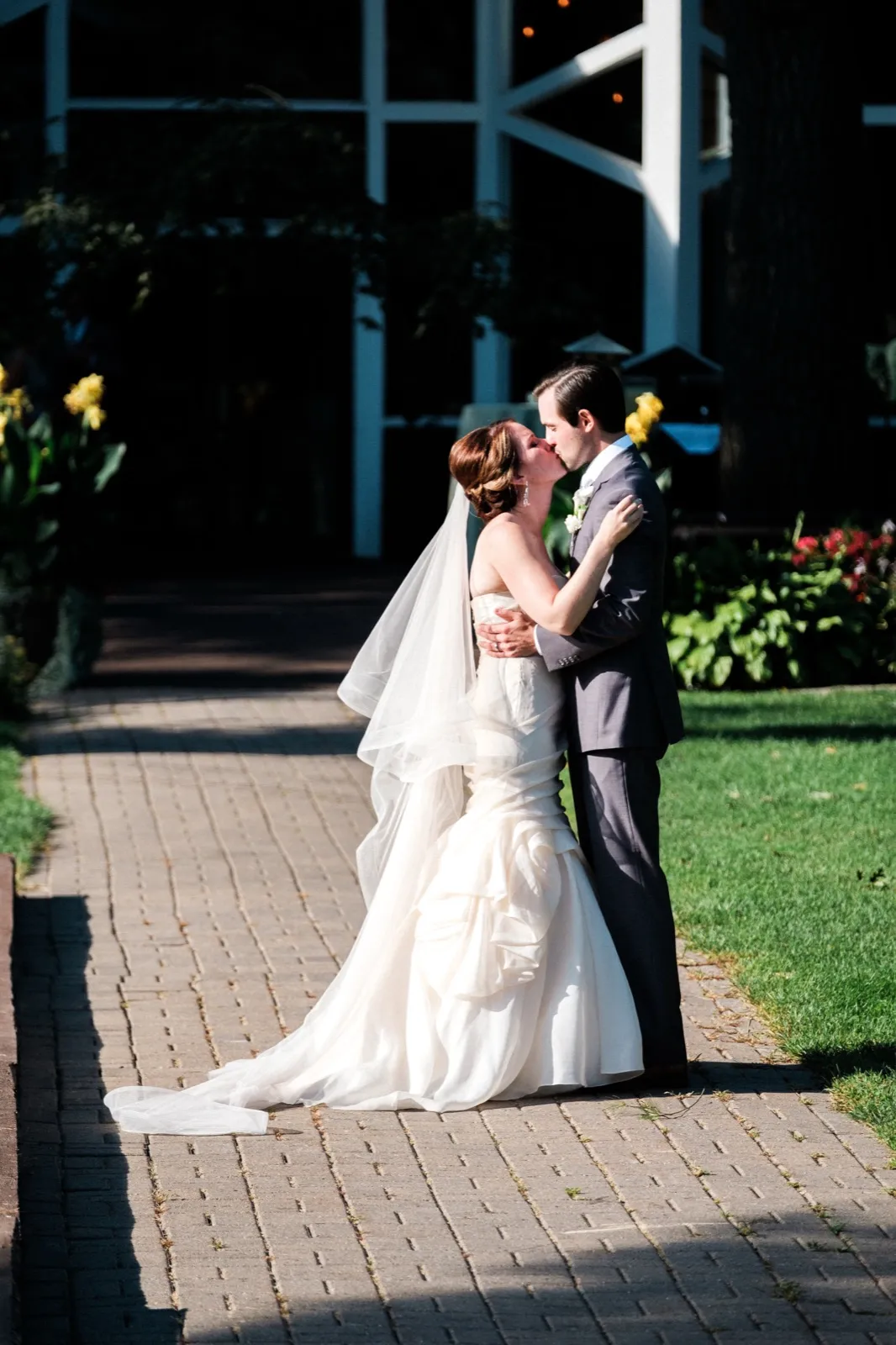 The bride and groom kiss on a sunlit brick pathway in front of the Grand View Lodge entrance, her veil and gown trailing behind her. — Tim Larsen Photography, Brainerd Lakes MN
