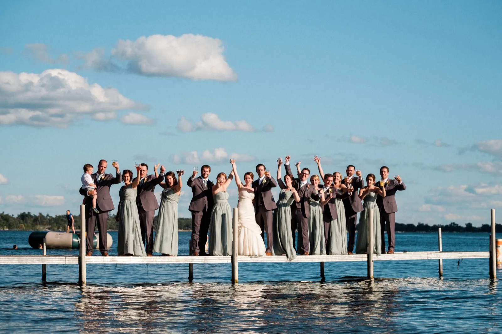 The bride, groom, and full bridal party stand in a line across a long lake dock at Grand View Lodge, raising drinks and cheering under a wide blue sky. — Tim Larsen Photography, Brainerd Lakes MN