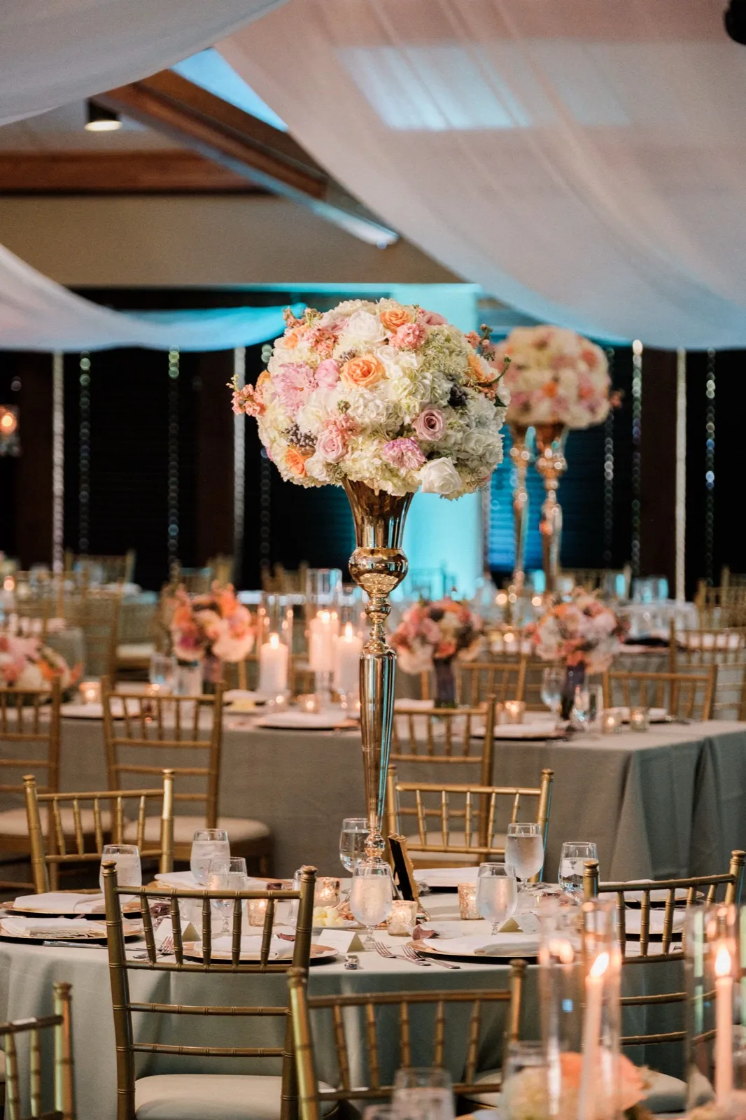 A tall gold centerpiece overflowing with pastel roses and hydrangeas rises above candlelit tables and gold chiavari chairs beneath the draped ceiling of the Grand View Lodge ballroom. — Tim Larsen Photography, Brainerd Lakes MN