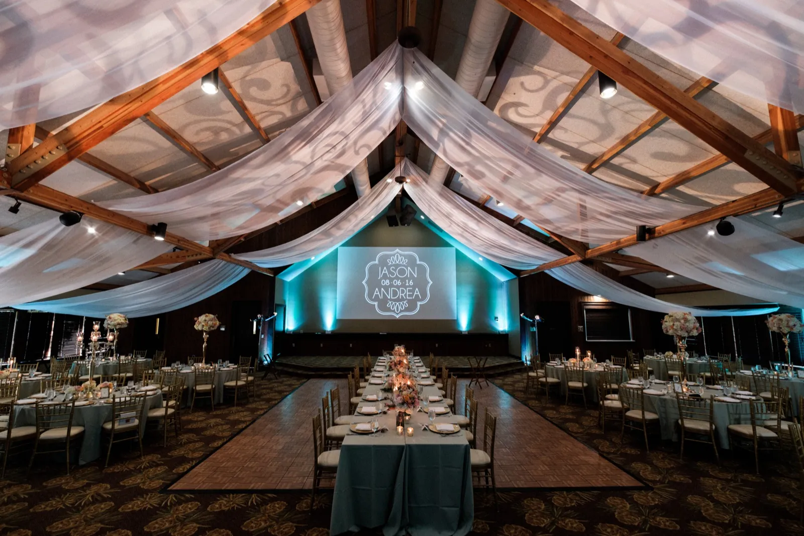 Wide symmetrical view of the Grand View Lodge ballroom set for dinner with draped ceiling fabric, exposed wood beams, and a 'Jason & Andrea 08·06·16' monogram projected on the back wall in teal light. — Tim Larsen Photography, Brainerd Lakes MN
