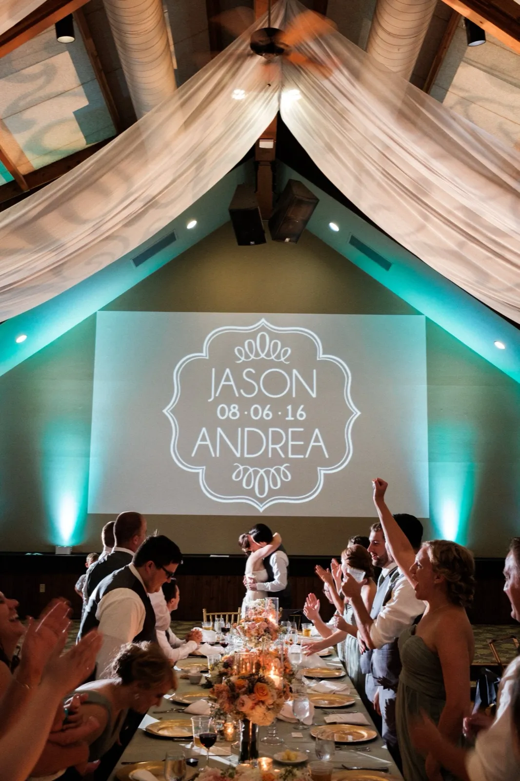 The couple kisses beside their head table as the bridal party cheers under Grand View Lodge's vaulted ballroom ceiling, draped in fabric and lit with teal uplighting beneath a projected JASON 08·06·16 ANDREA monogram. — Tim Larsen Photography, Brainerd Lakes MN
