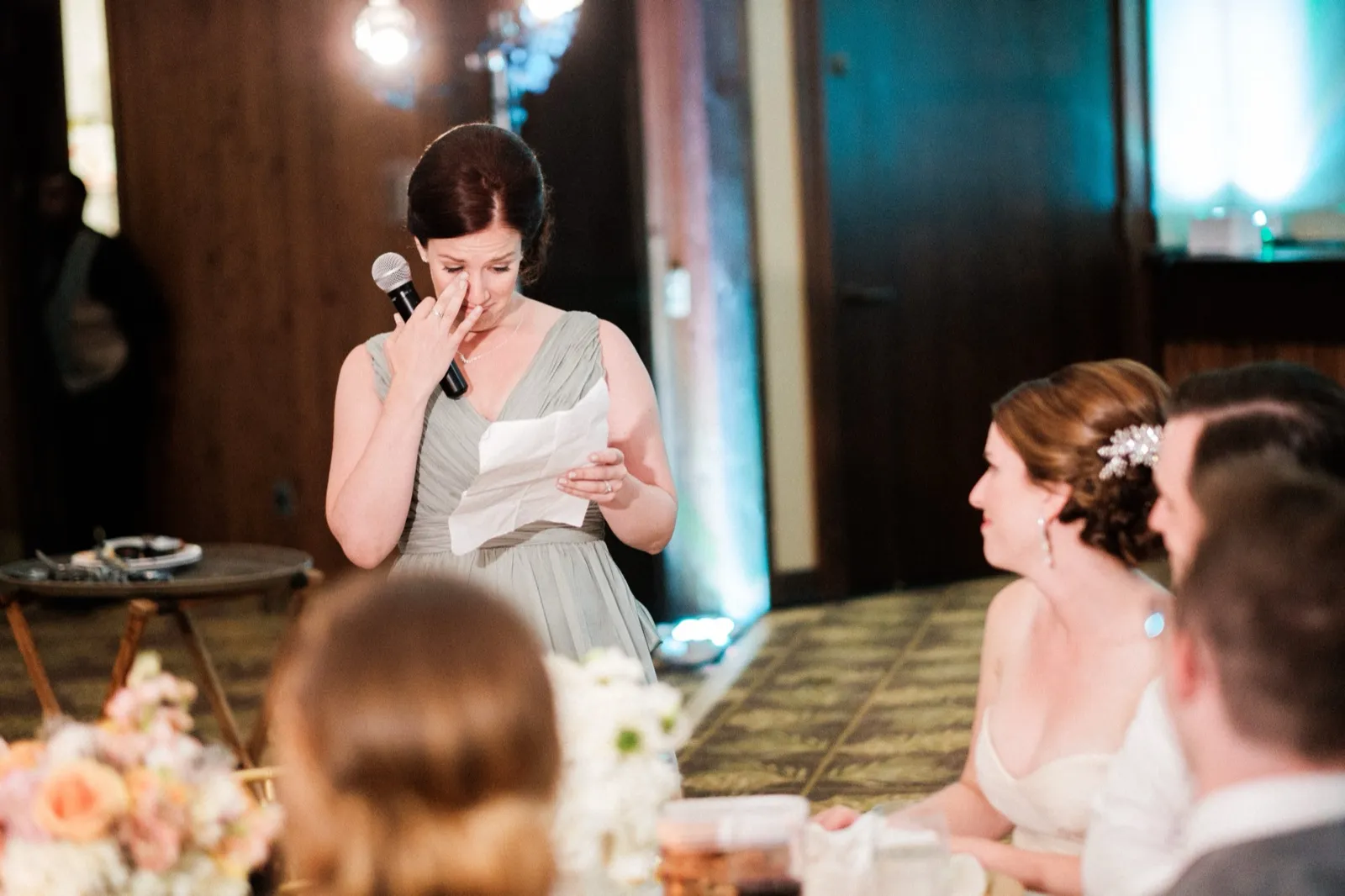 A bridesmaid in a sage green dress holds a microphone and wipes a tear from her eye while reading from a folded paper, as the bride watches from the foreground. — Tim Larsen Photography, Brainerd Lakes MN