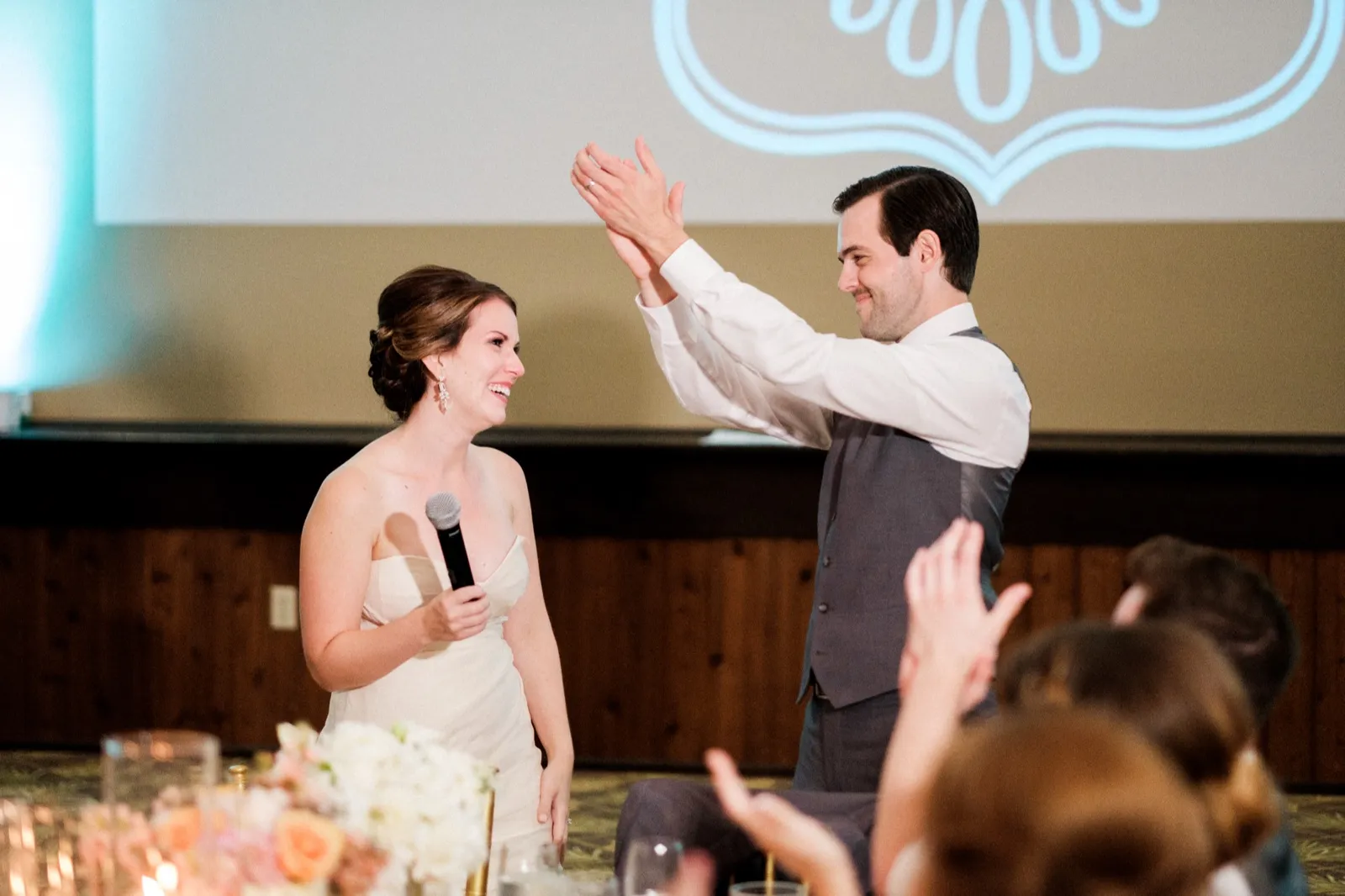 The bride holds a microphone and laughs as the groom raises his hand for a high-five during reception toasts, with applauding guests in the foreground. — Tim Larsen Photography, Brainerd Lakes MN