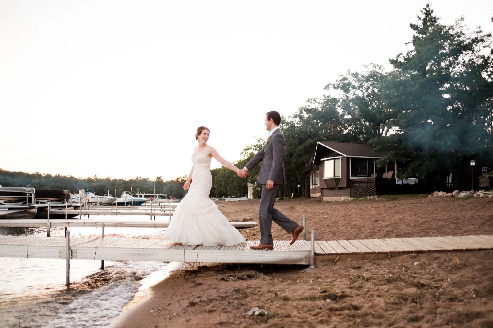 The bride laughs as she leads the groom by the hand across a wooden dock at dusk, with pontoon boats moored to the left and a lakeshore cabin behind them. — Tim Larsen Photography, Brainerd Lakes MN