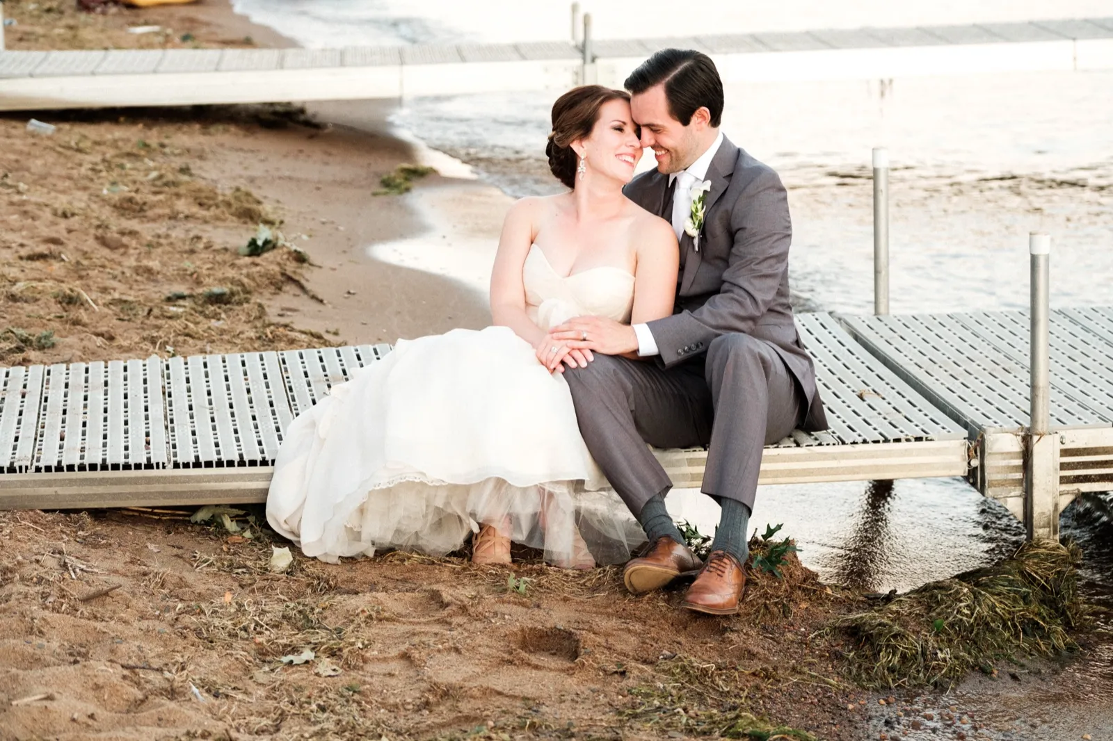 The bride and groom sit together on the edge of a lakeside dock at Grand View Lodge, their foreheads touching as she laughs softly. — Tim Larsen Photography, Brainerd Lakes MN