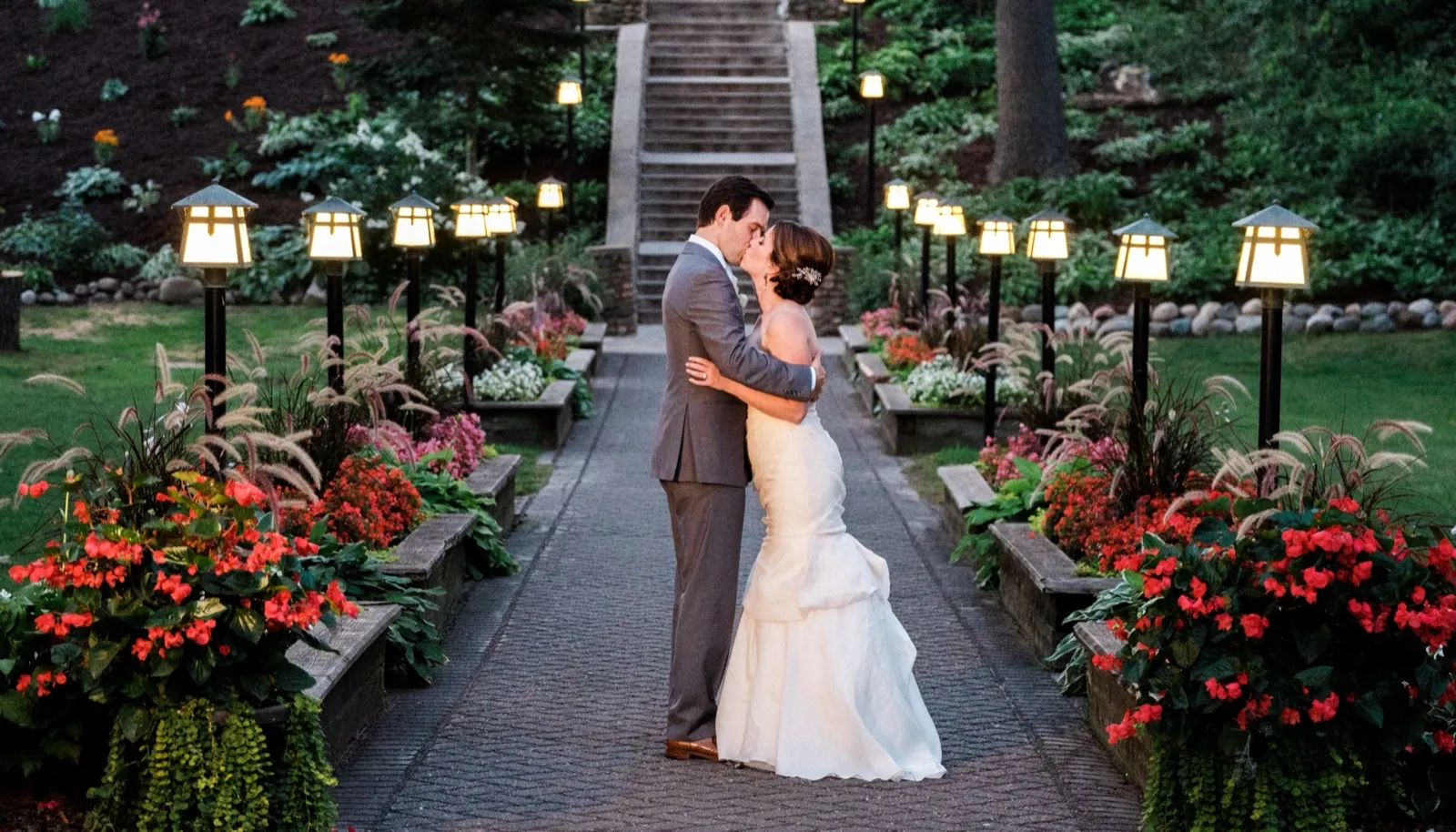 The bride and groom kiss on a brick pathway lined with glowing craftsman lanterns and red flower beds at dusk, with a stone stairway rising behind them. — Tim Larsen Photography, Brainerd Lakes MN