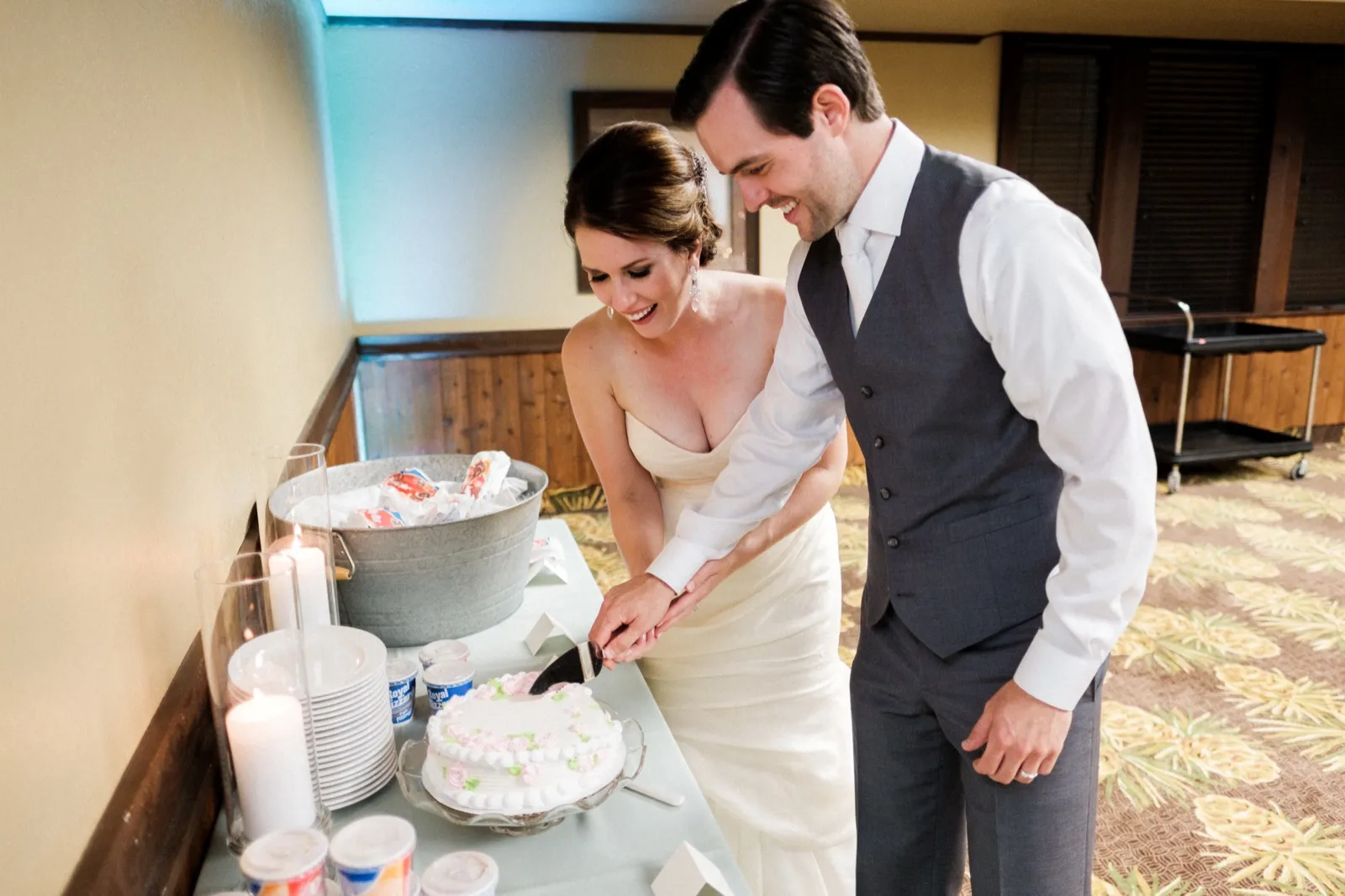 The bride and groom lean together laughing as they cut a small rose-decorated cake at a reception side table set with candles and a tub of ice cream. — Tim Larsen Photography, Brainerd Lakes MN