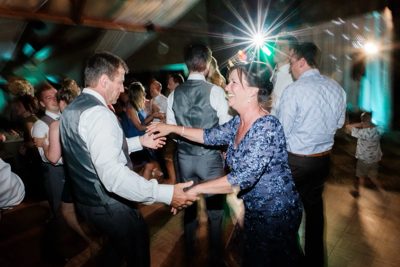 A man and a woman in a navy patterned dress laugh while clasping hands and dancing under a starburst stage light surrounded by other guests. — Tim Larsen Photography, Brainerd Lakes MN