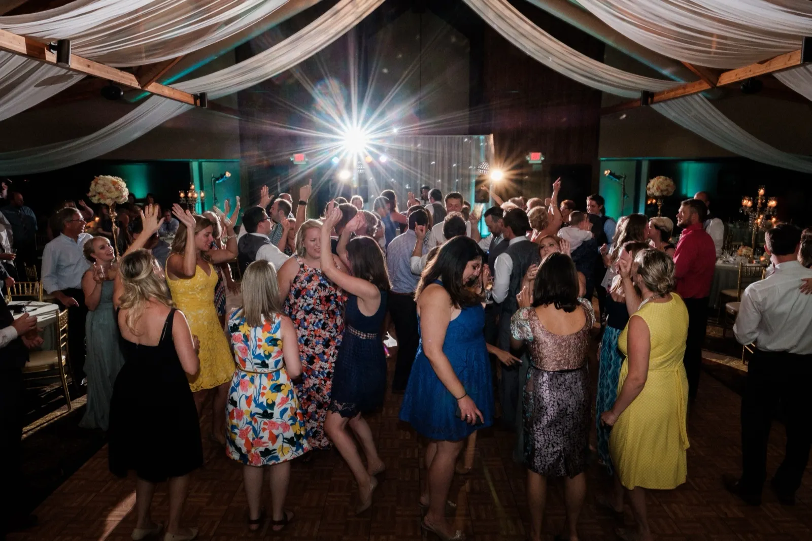A packed indoor dance floor of wedding guests raises their hands beneath sweeping draped fabric and a bright starburst stage light at Grand View Lodge. — Tim Larsen Photography, Brainerd Lakes MN