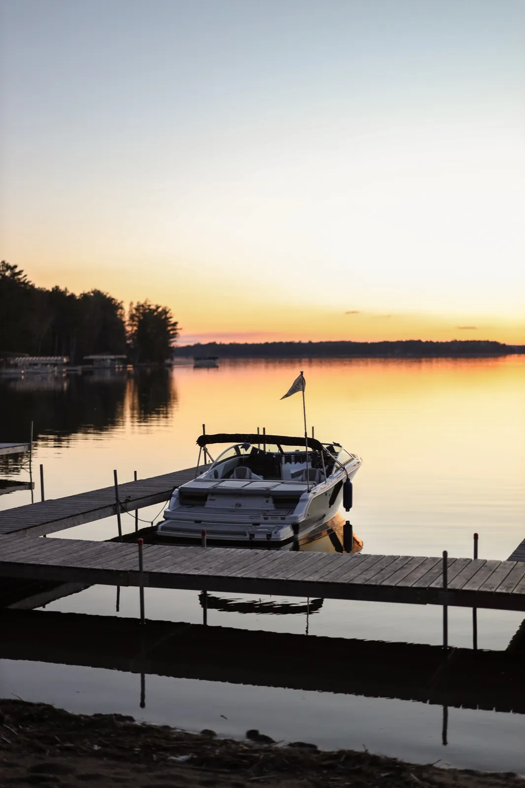 A small boat with a flag is moored to a wooden dock on a still lake at dusk, glowing horizon and mirrored reflections in the water — Tim Larsen Photography, Brainerd Lakes MN