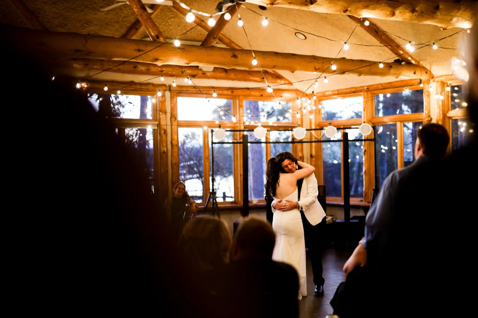The bride and groom embrace during their first dance beneath a canopy of string lights and exposed log beams, framed by silhouetted guests watching from the edges of the room — Tim Larsen Photography, Brainerd Lakes MN