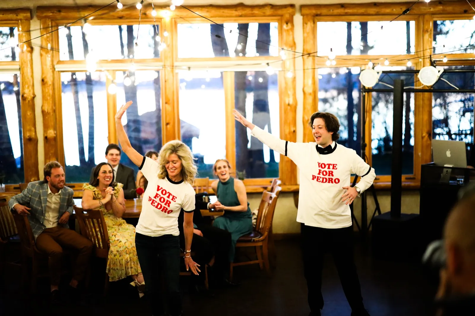 Two guests in matching 'Vote for Pedro' shirts perform a synchronized dance while seated guests laugh and watch behind them at the lakeside lodge reception — Tim Larsen Photography, Brainerd Lakes MN