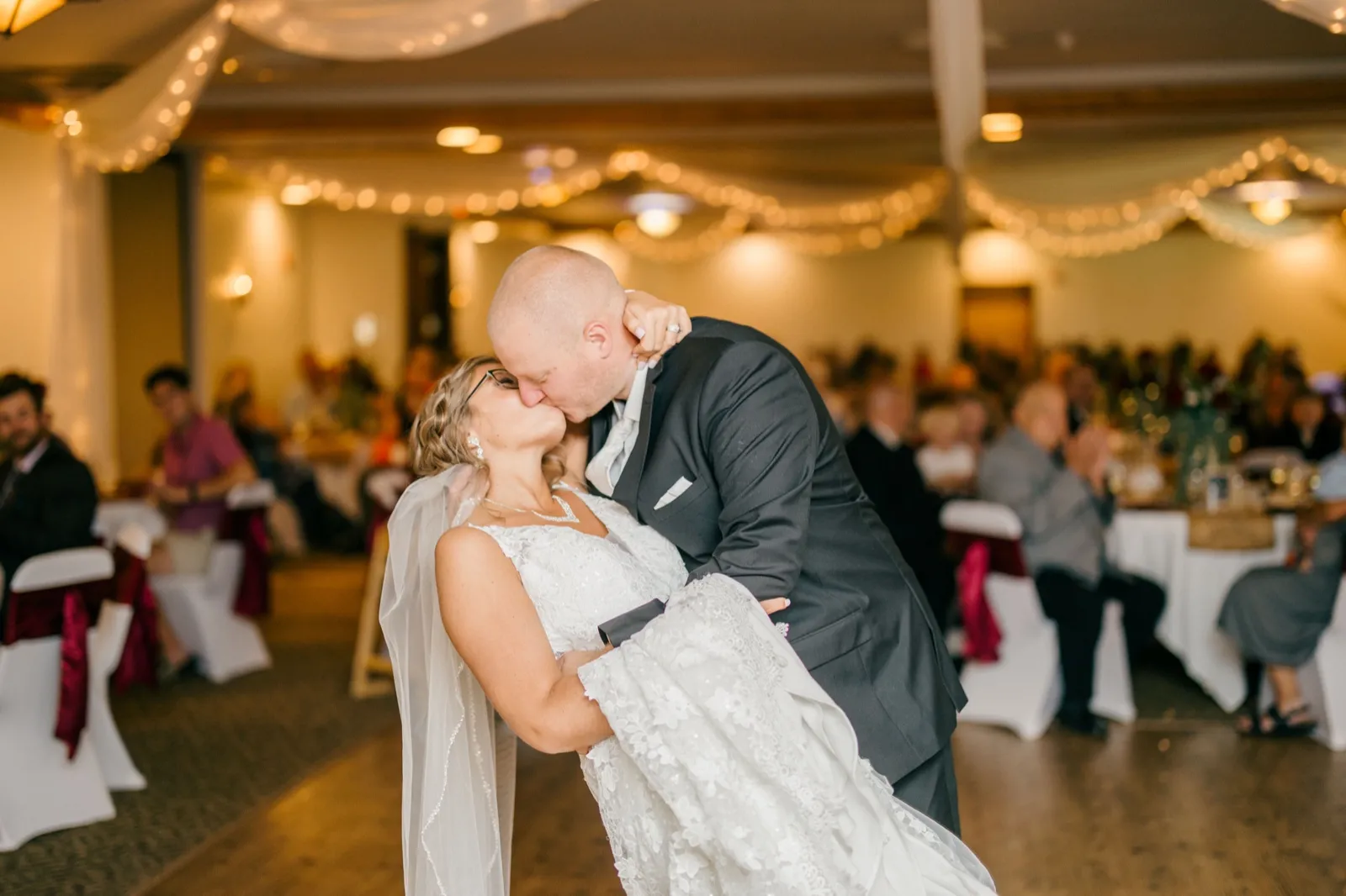Kim and Jesse kiss on the dance floor with her veil sweeping behind them, beneath draped fabric and string lights with seated guests visible in the background — Tim Larsen Photography, Brainerd Lakes MN