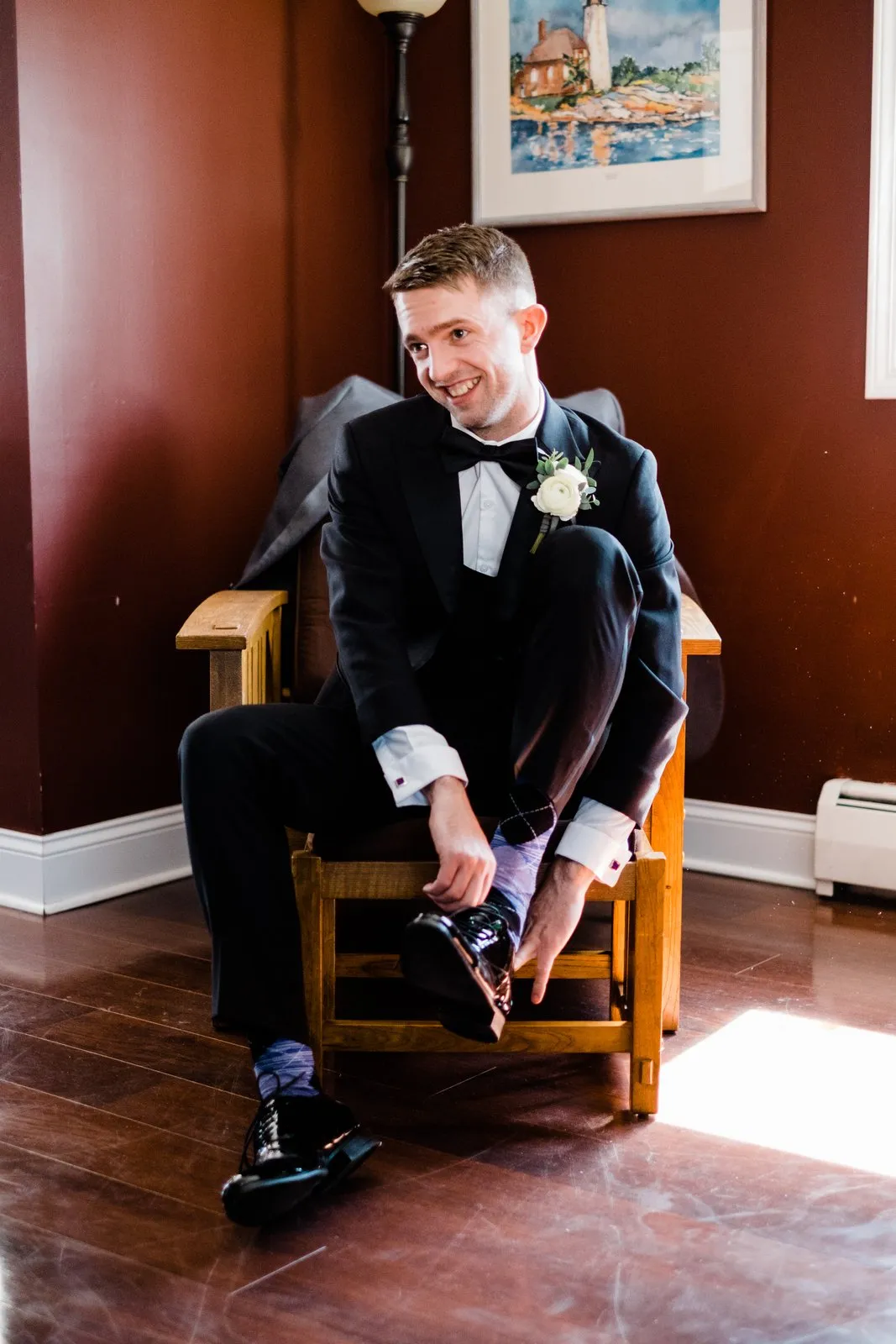 Groom mid-grin in a mission chair, lighthouse painting overhead — Duluth Catholic spring wedding getting-ready portrait — Tim Larsen Photography, Duluth MN