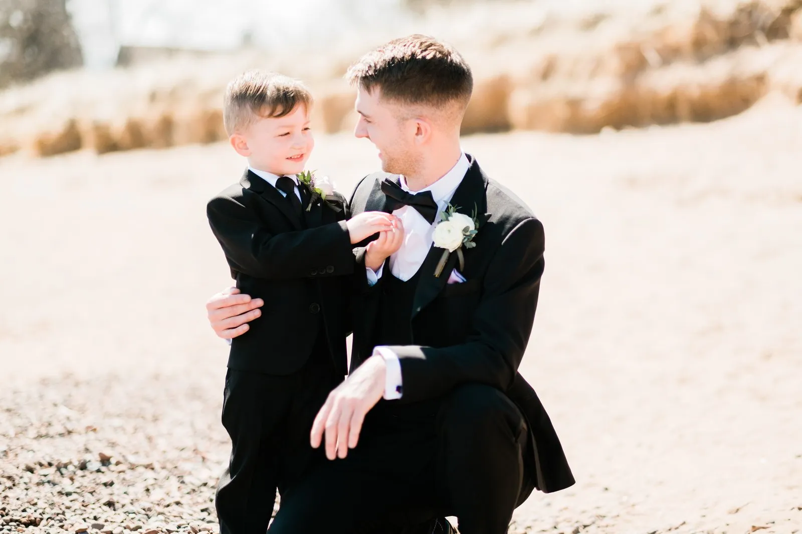 Groom kneels beside the young ring bearer on the Lake Superior shoreline, dune grass behind — Duluth Catholic spring wedding portrait — Tim Larsen Photography, Duluth MN