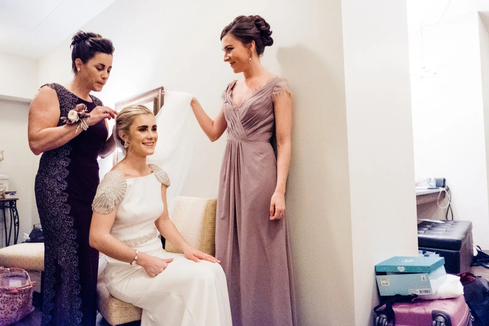 Bride seated as her mother and a bridesmaid arrange her cathedral veil, framed against pale wall — Greysolon Plaza bridal suite — Tim Larsen Photography, Duluth MN