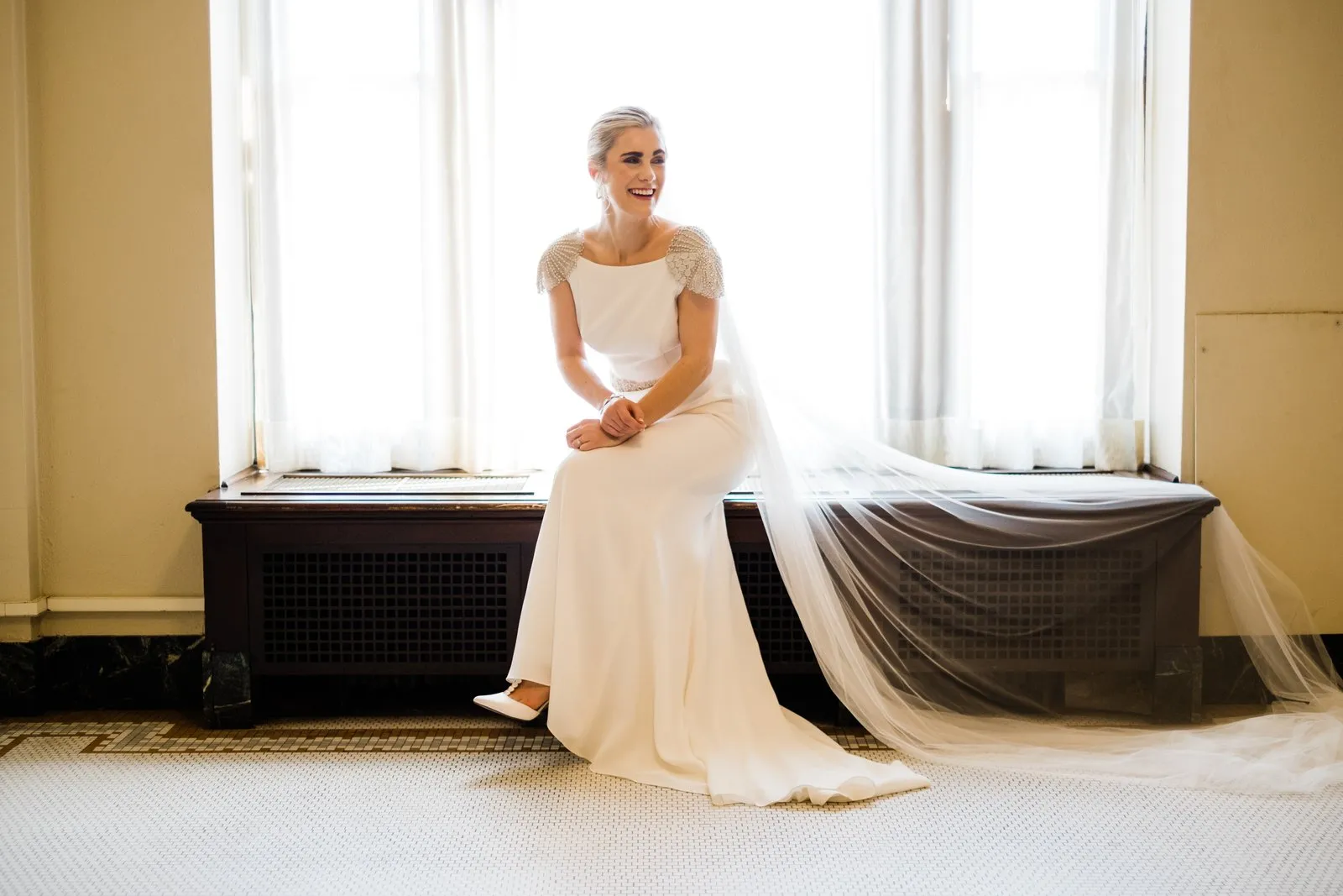 Bride seated between tall windows, cathedral veil cascading across the bench — Greysolon Plaza bridal suite, Duluth Catholic wedding — Tim Larsen Photography, Duluth MN