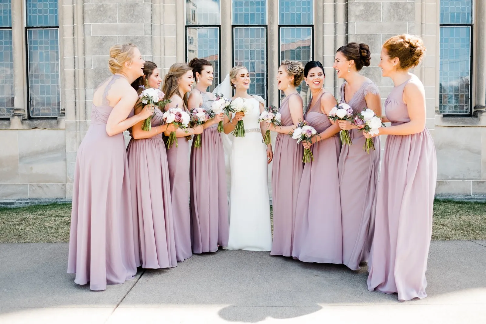Bride and bridesmaids in dusty lavender arc inward against the stained-glass gothic facade of the Cathedral of Our Lady of the Rosary — Duluth Catholic wedding — Tim Larsen Photography, Duluth MN