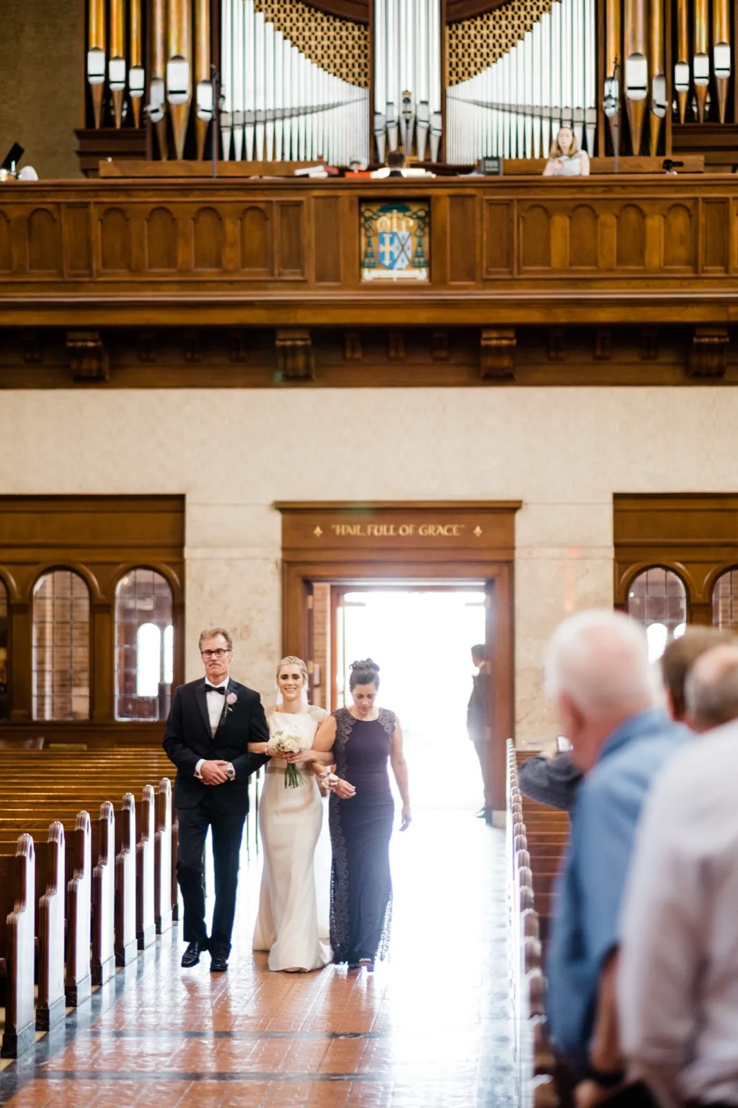 Symmetrical view down the aisle — bride backlit by the open doorway, ornate organ pipes above — Cathedral of Our Lady of the Rosary processional — Tim Larsen Photography, Duluth MN