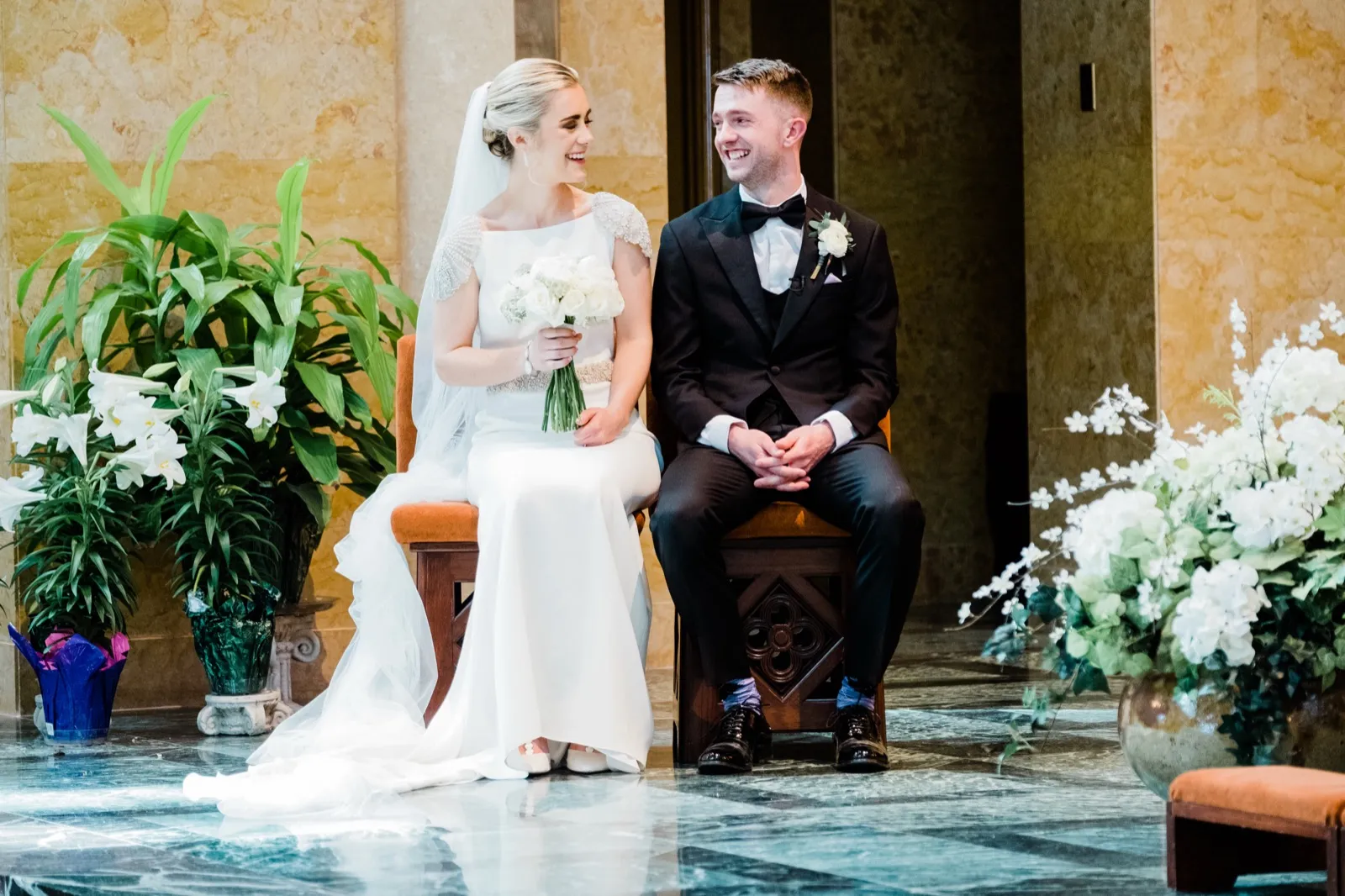 Symmetrical seated couple framed by Easter lilies and marble pillars during shared laughter mid-ceremony — Cathedral of Our Lady of the Rosary — Tim Larsen Photography, Duluth MN