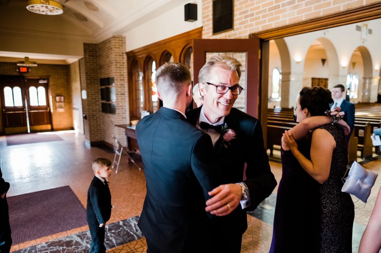 Father-son embrace at the center as the mother hugs a guest at right — tender family candid, Cathedral of Our Lady of the Rosary Duluth — Tim Larsen Photography, Duluth MN