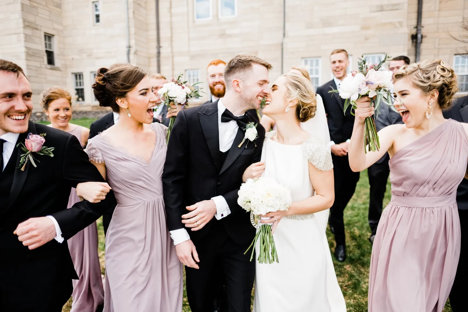 Couple kiss center-frame as the bridal party erupts with raised bouquets and open laughter — outside the Cathedral, Duluth Catholic spring wedding — Tim Larsen Photography, Duluth MN