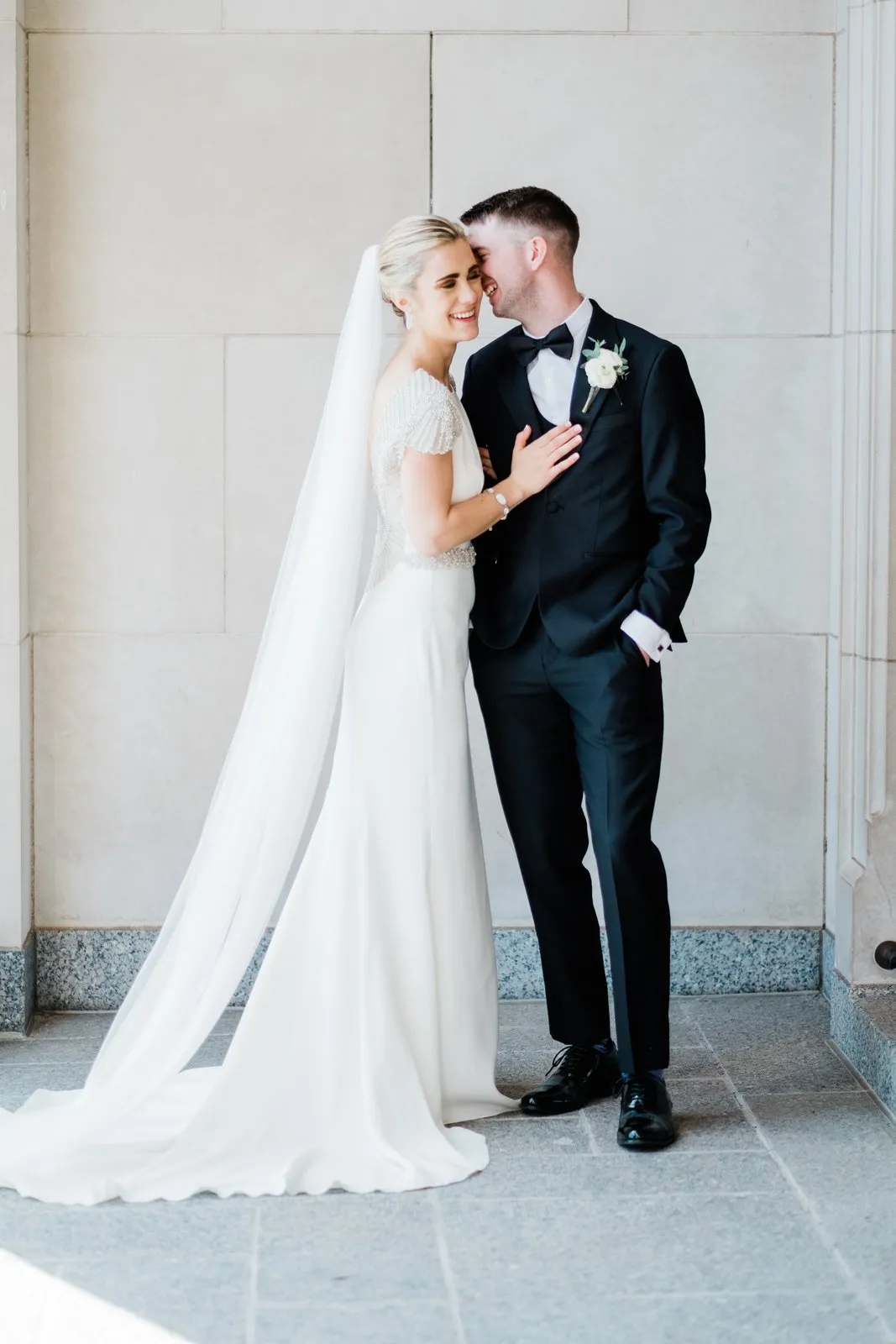 Full-length couples portrait against a limestone block wall, cathedral veil pooling left — downtown Duluth Catholic spring wedding — Tim Larsen Photography, Duluth MN