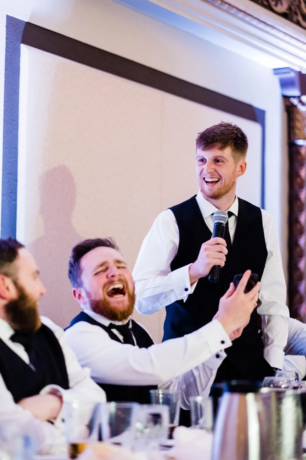 Speaker mid-toast as a seated groomsman erupts in head-back laughter beside him — Greysolon Ballroom reception, Duluth wedding — Tim Larsen Photography, Duluth MN
