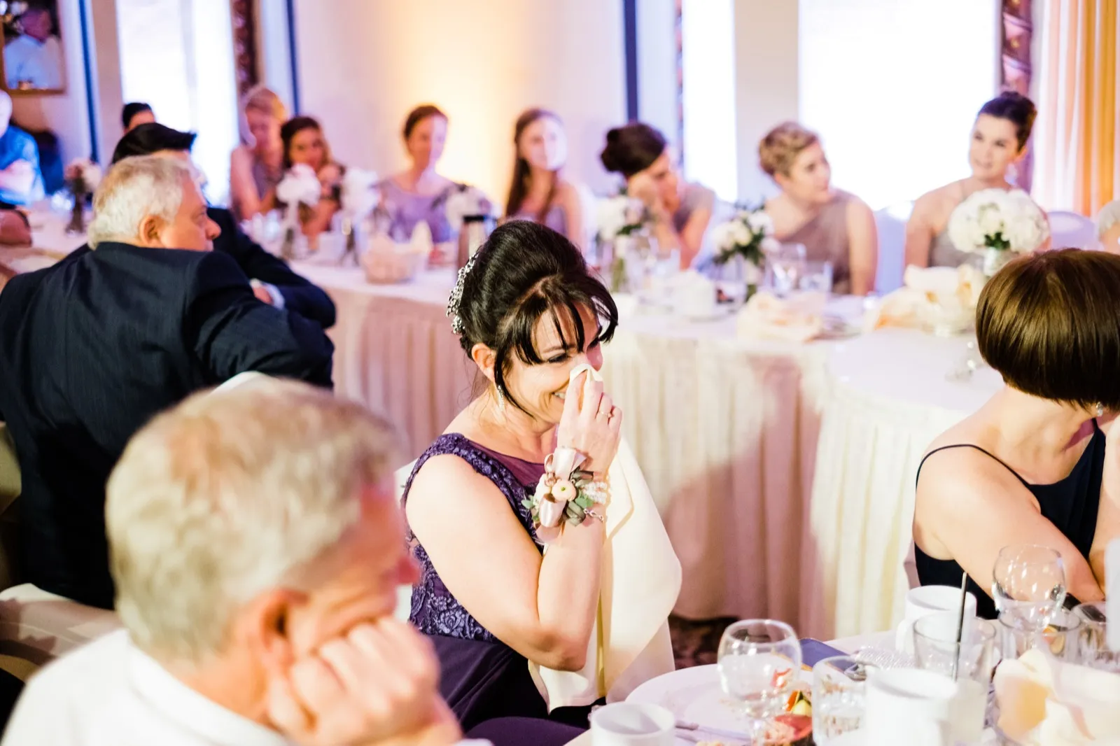 Mother dabs tears mid-toast, framed between two foreground guests at adjacent tables — Greysolon Ballroom reception, Duluth Catholic wedding — Tim Larsen Photography, Duluth MN
