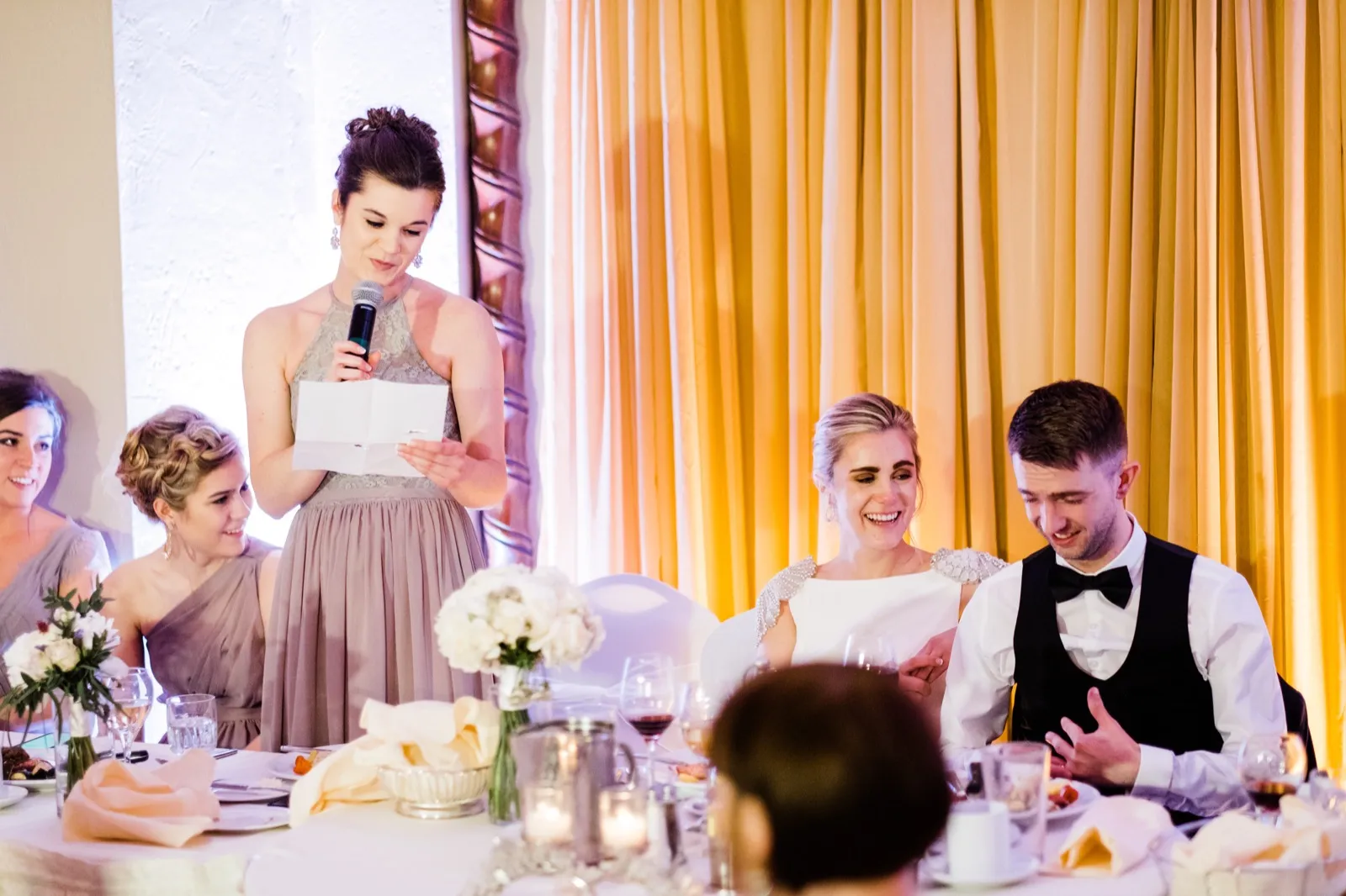 Bridesmaid reads from a card at left as the couple laughs warmly at right against gold drapery — Greysolon Ballroom reception, Duluth wedding — Tim Larsen Photography, Duluth MN