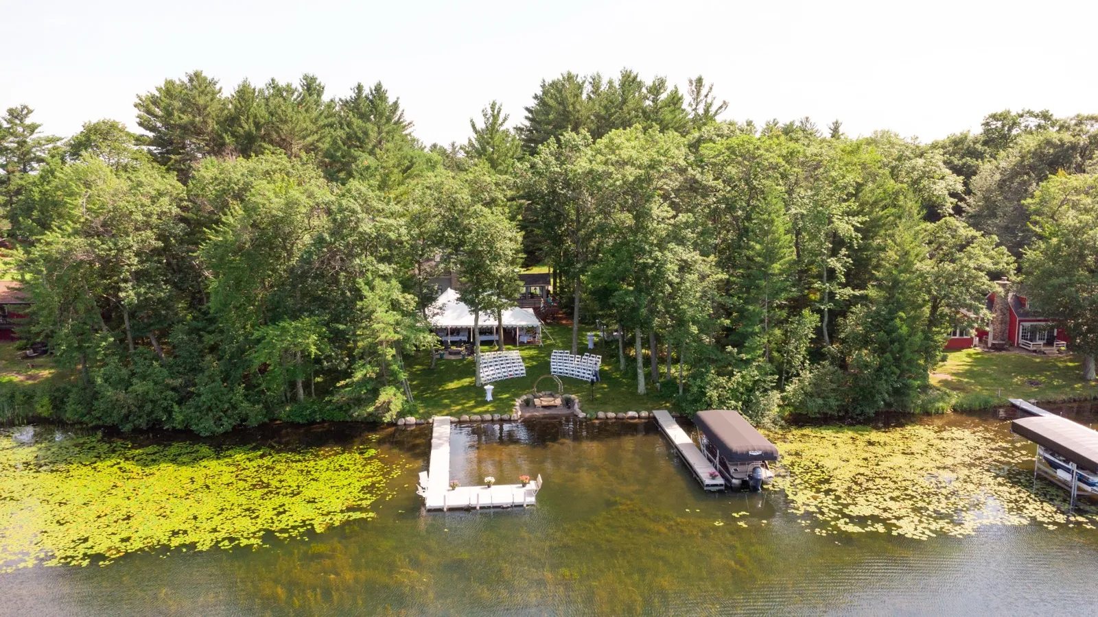 An aerial view of a private lakeside cabin ceremony site with a white tent, rows of chairs facing an arch, a dock, and a pontoon boat on lily-pad-dotted water. — Tim Larsen Photography, Brainerd Lakes MN