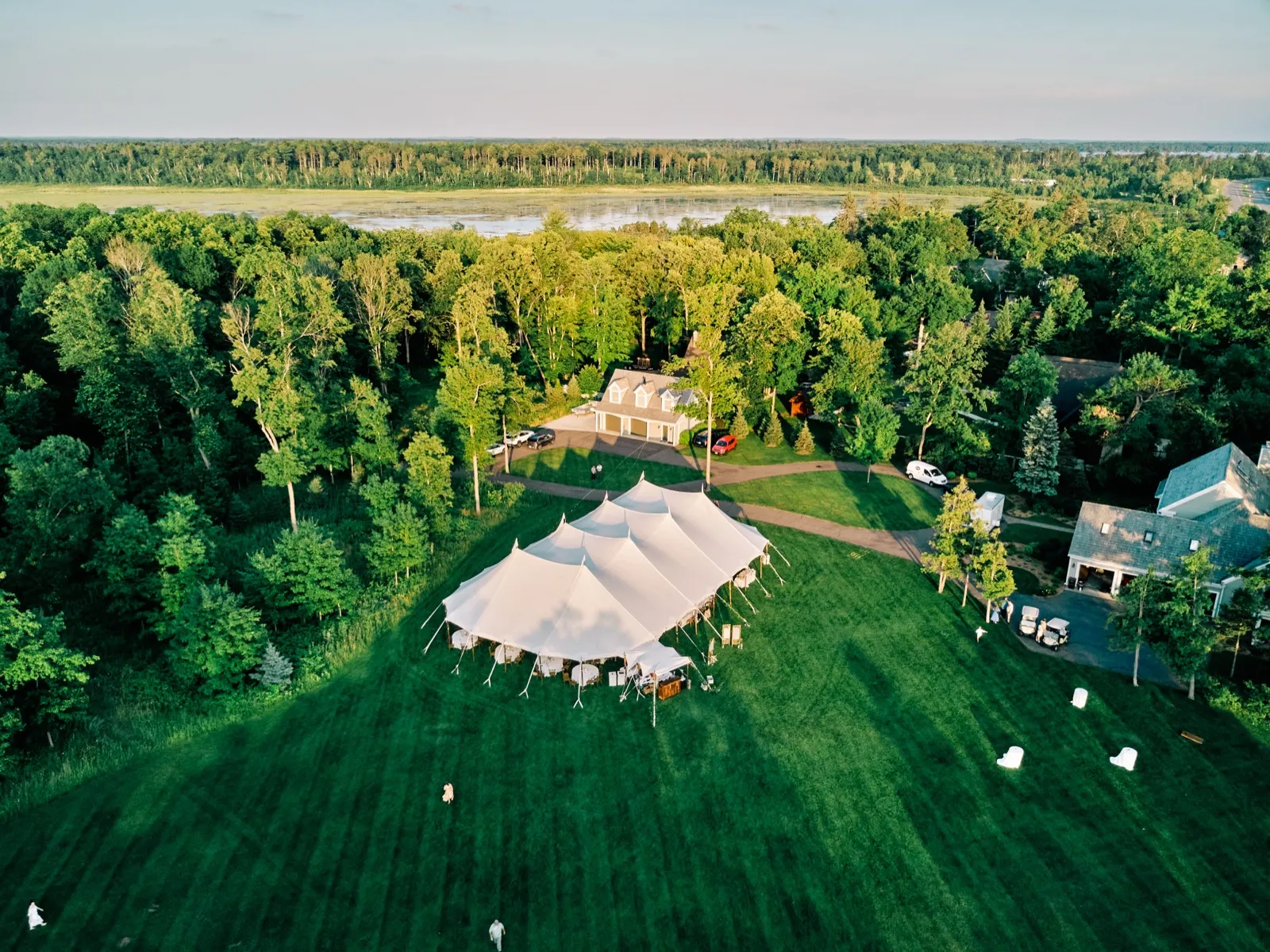 Aerial view of a large white sailcloth reception tent set on a sprawling green lawn beside a private lake cabin, with forest and a lake stretching to the horizon at golden hour. — Tim Larsen Photography, Brainerd Lakes MN