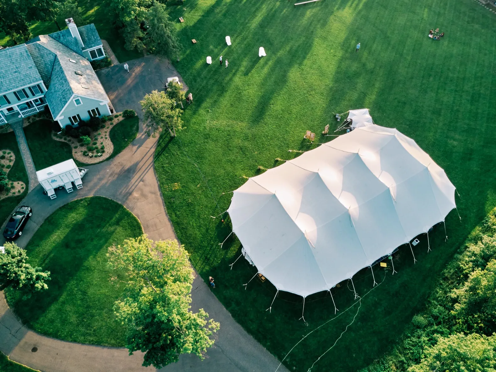 Aerial view of a large white pole tent set up on a sprawling green lawn beside a lake house, with guests and vendors moving across the grass in afternoon light. — Tim Larsen Photography, Brainerd Lakes MN