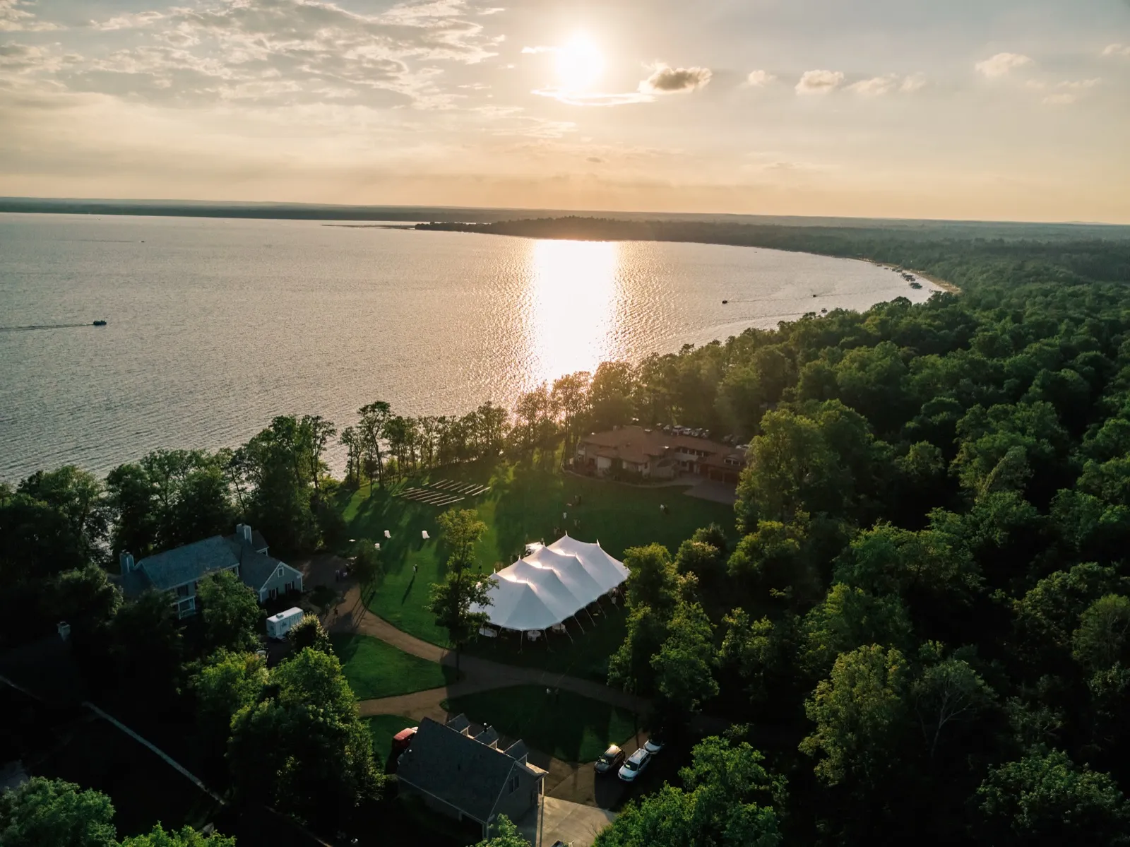 An aerial view of a lakeside cabin property on Gull Lake at golden hour, with a white sailcloth reception tent on the lawn and the sun reflecting across the water. — Tim Larsen Photography, Brainerd Lakes MN