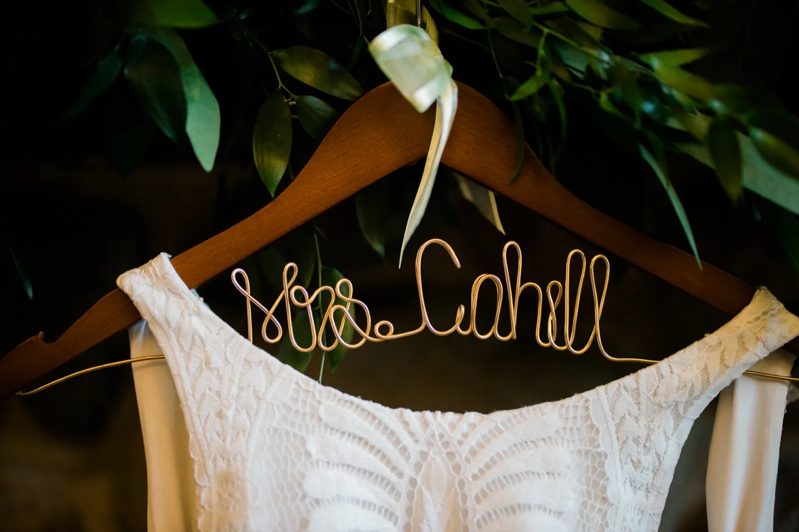 A custom wire hanger reading 'Mrs. Cahill' holds the bride's lace wedding dress beneath a draping of green leaves. — Tim Larsen Photography, Brainerd Lakes MN