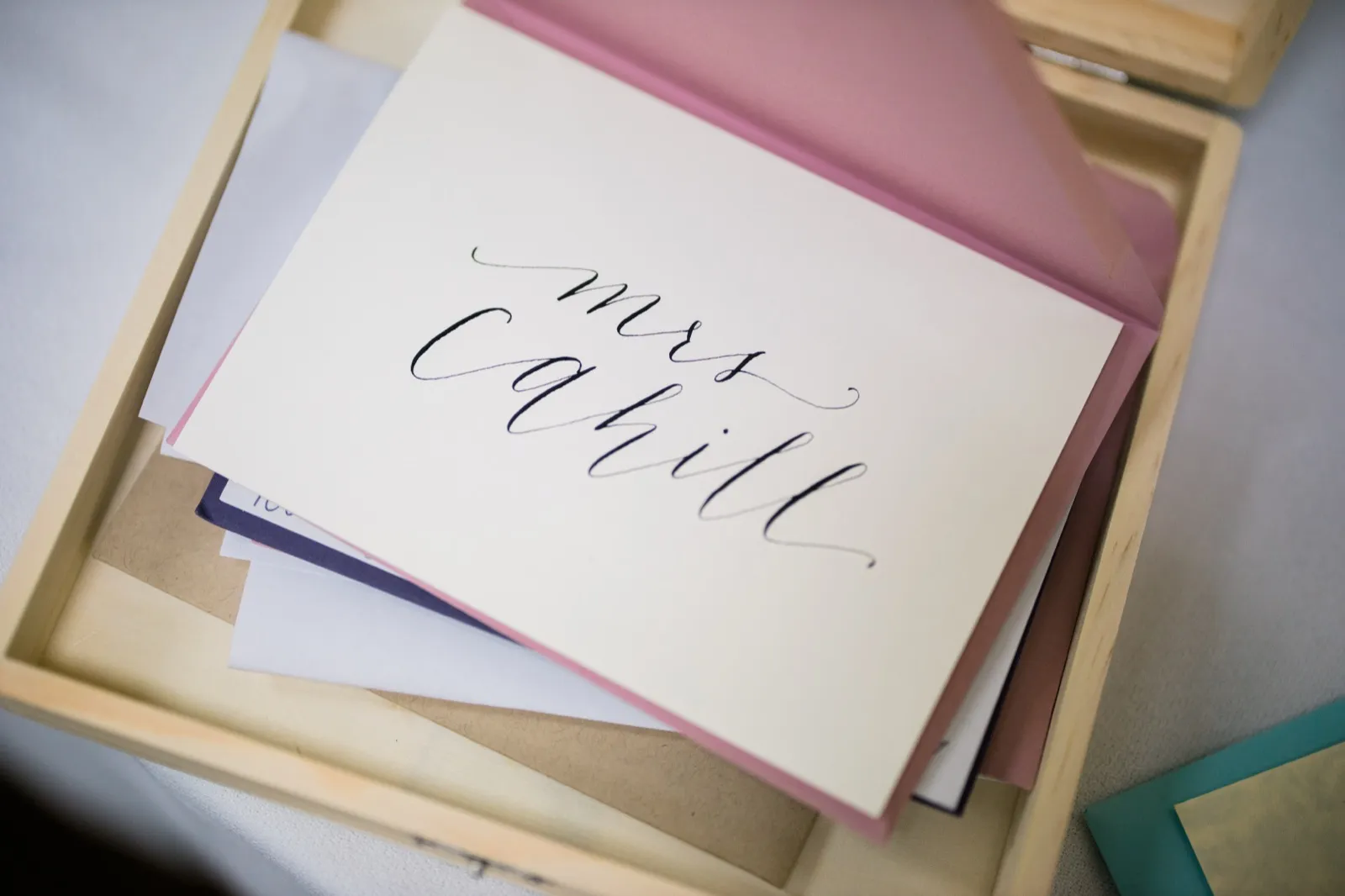 A handwritten calligraphy card reading 'Mrs Cahill' rests on a dusty rose envelope inside a wooden tray of stationery. — Tim Larsen Photography, Brainerd Lakes MN